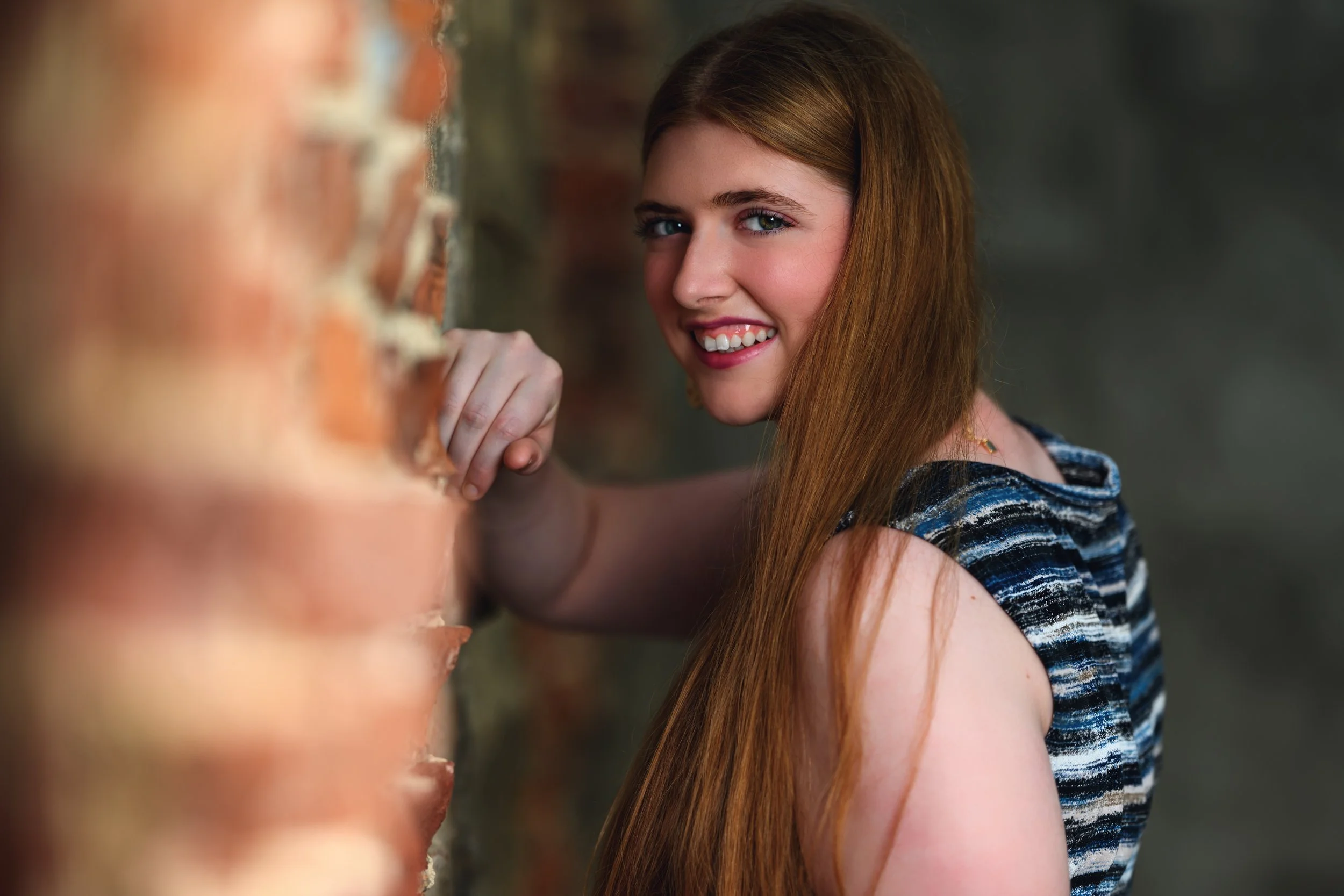 A young woman with long red hair and blue eyes smiling and looking at the camera, leaning on a brick wall.