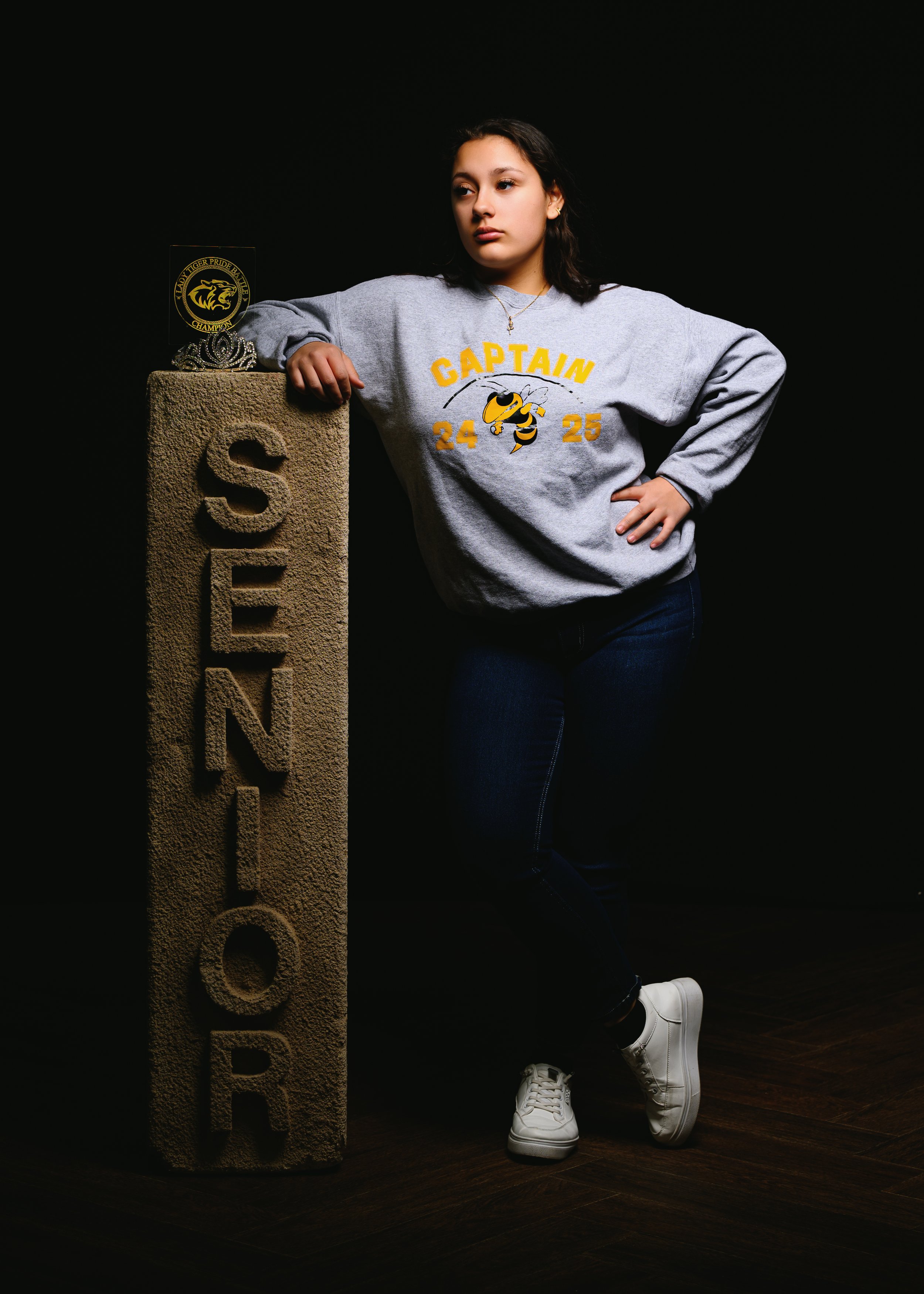 A person stands leaning against a stone pillar with the word "SENIOR" engraved on it. They are wearing a gray sweatshirt with "CAPTAIN 24 25" and a bee design, along with jeans and white sneakers. A small trophy is also placed on top of the stone pil