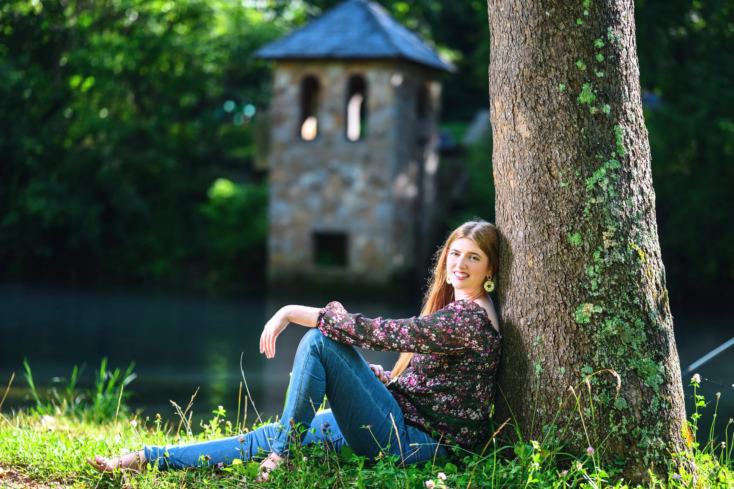 A young woman with long hair, wearing a floral blouse and blue jeans, sitting on the grass leaning against a tree trunk by a body of water, with a stone tower in the background.