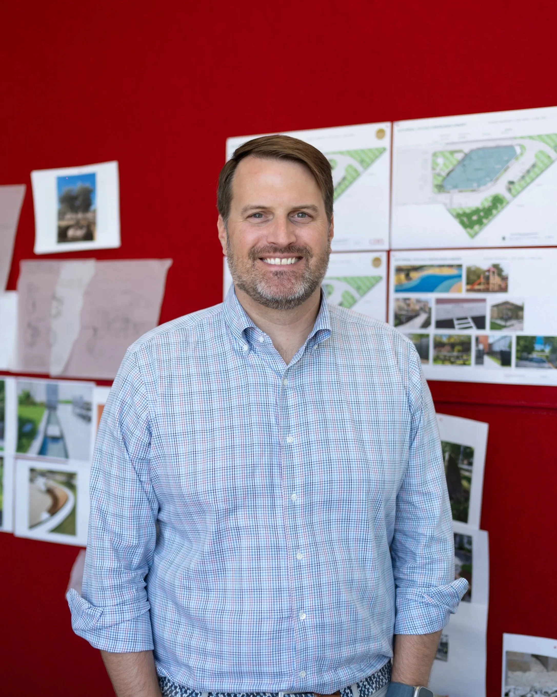 A man with light brown hair, a beard, and a checkered light blue shirt smiling in front of a red wall with architectural plans and photos pinned to it.