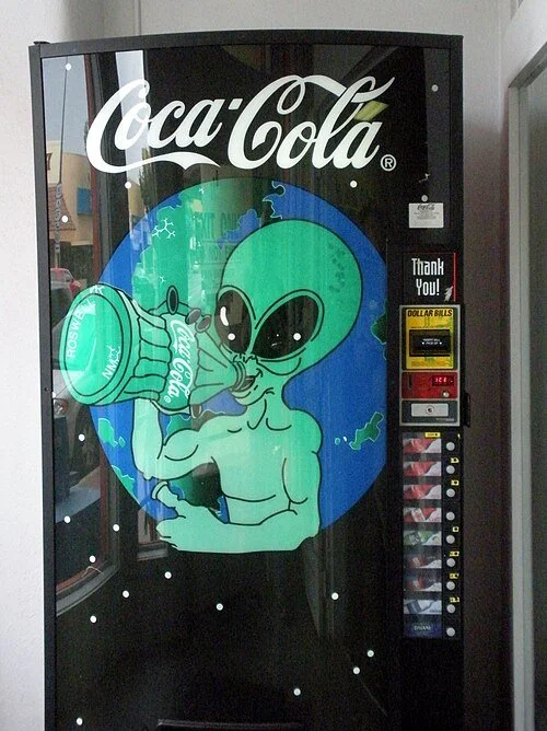 Photo of a Coca-Cola vending machine bearing the image of a green extraterrestrial drinking a bottle of Coke