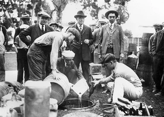 B&W photo of three men pouring liquor into a sewer while a group of other men look on; all are wearing 1920s-era clothing