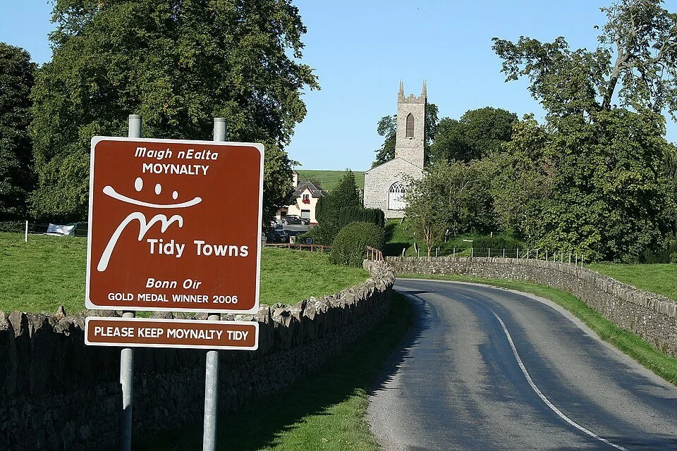 Country road leading into Moynalty with a sign saying it won the 2006 Tidy Towns competition; a church is in the background