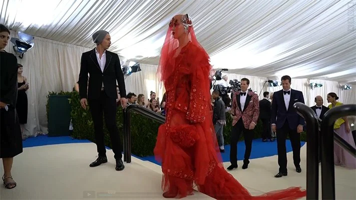 A woman in a haute couture, red dress with veil walking through a tented enclosure with people in evening wear following