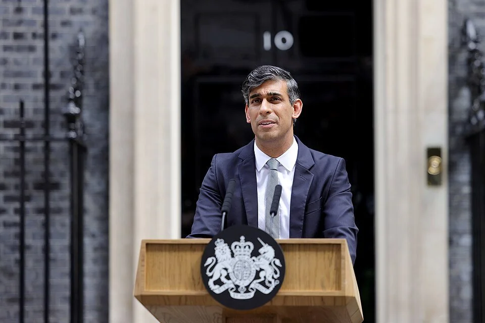 A desi man in a suit standing at a lectern in front of 10 Downing Street, London