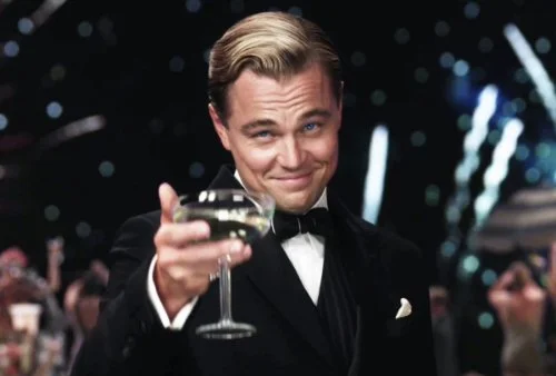 Screenshot of a smiling, tuxedo-clad Leonardo DiCaprio raising a glass of champagne in a toast