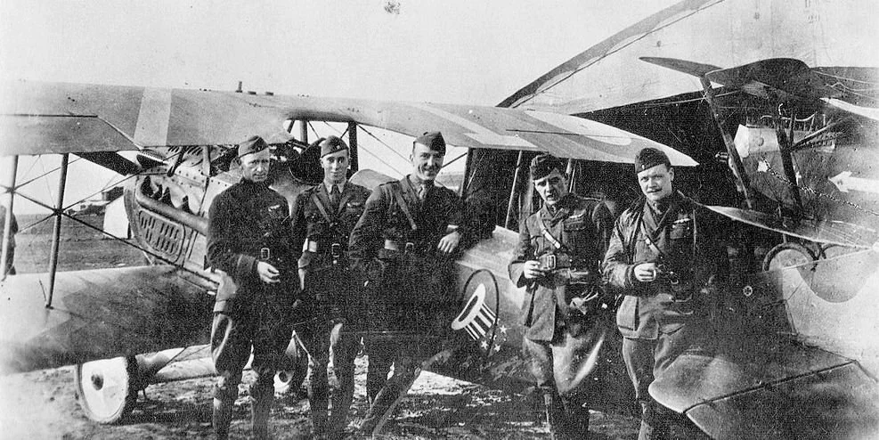 B&W photo of five pilots in front of a WWI aircraft bearing a squadron insignia of a stars-and-stripes top hat in a ring