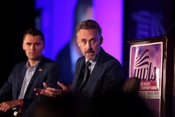 Photo of two men in suits seated on a stage, one gesturing and speaking to someone off camera