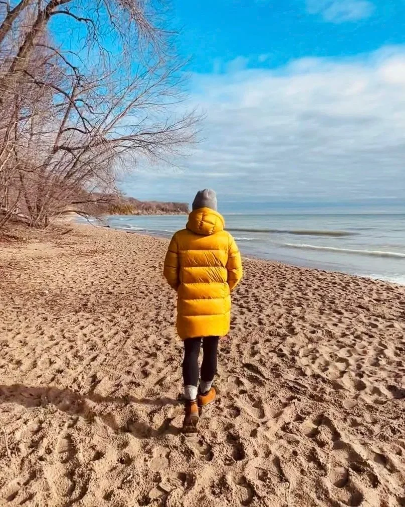 Tamara Okun Valerine in a yellow puffy jacket, gray beanie, and black pants walking on a sandy beach with bare trees on the left and the ocean on the right, under a partly cloudy blue sky.