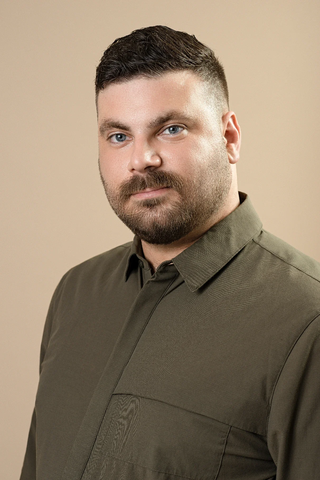 A man with short dark hair, blue eyes, and a beard wearing an olive green button-up shirt. He has a neutral expression and is facing slightly to the left against a plain beige background.