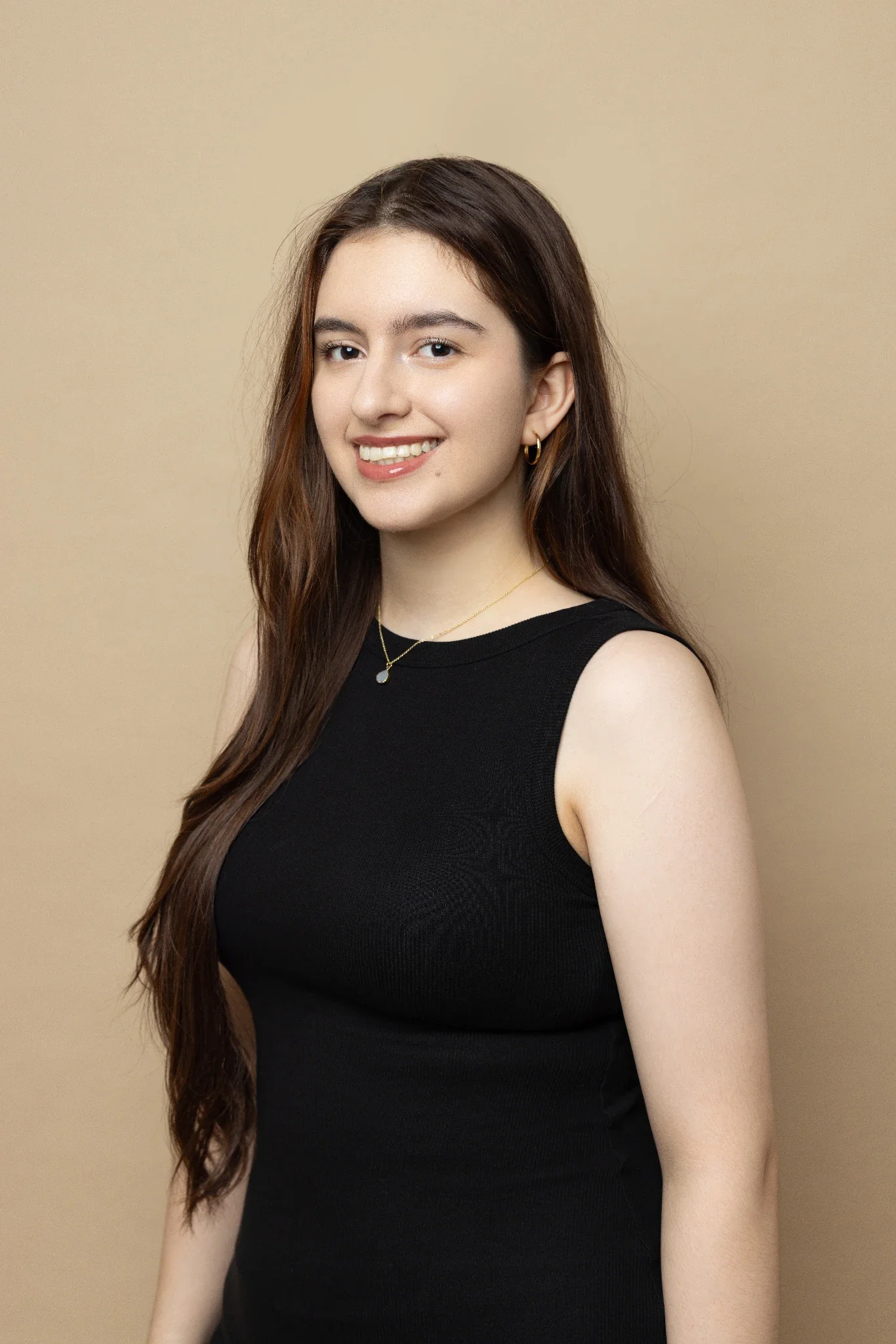 A young woman with long wavy brown hair, winking and smiling, wearing a black sleeveless top, jewelry, and standing against a beige background.