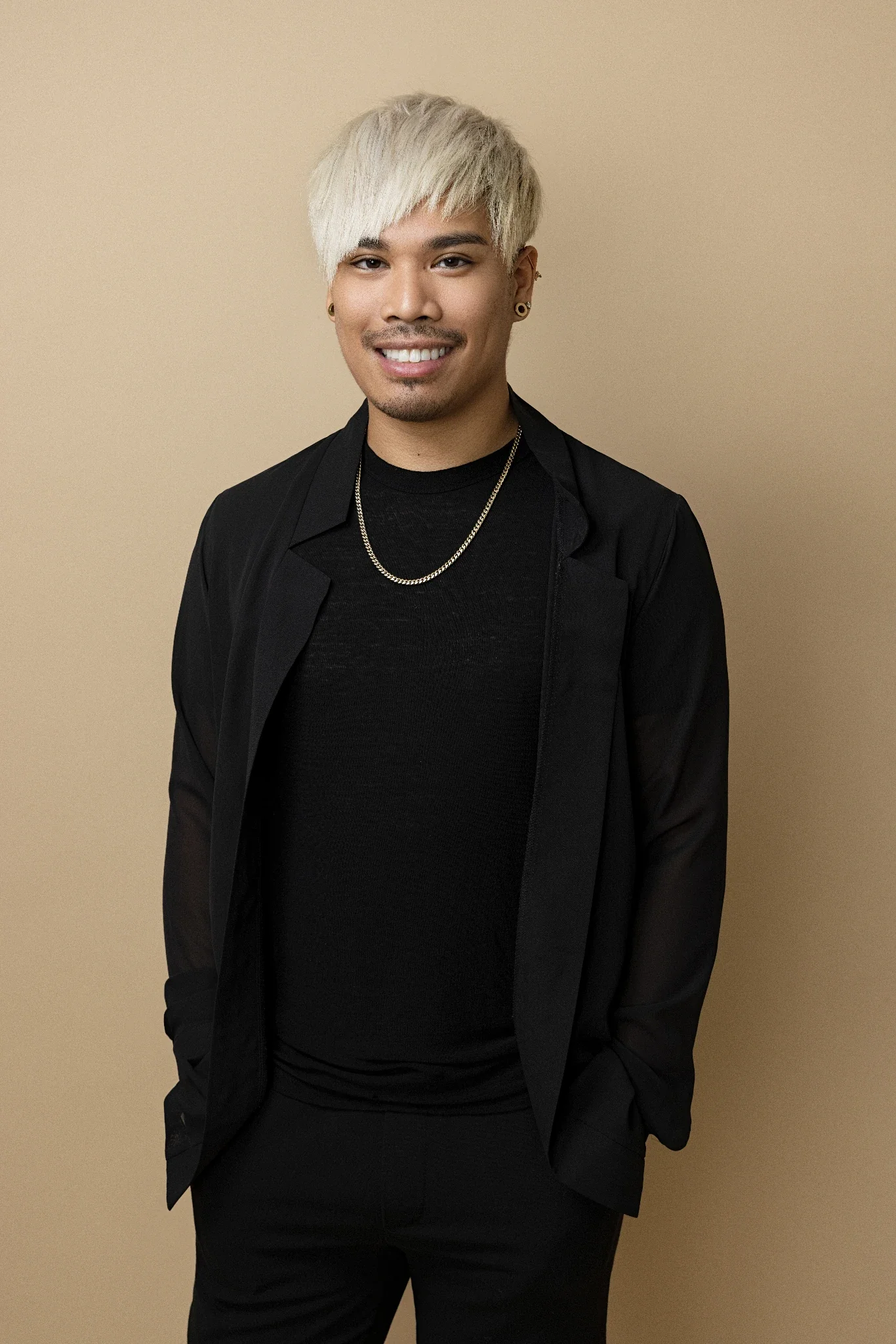 Portrait of a young man with platinum blonde hair, wearing a black blazer, black shirt, gold chain, and earrings, standing against a beige background.