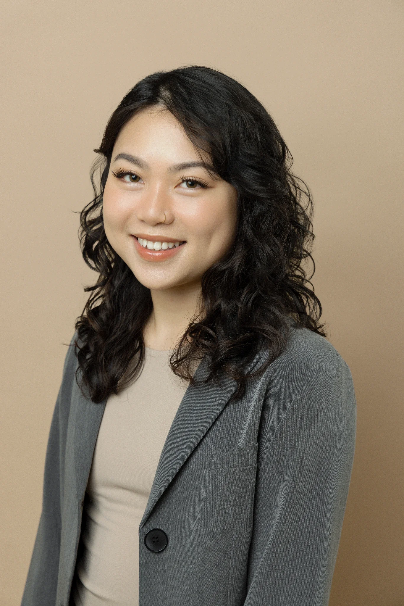 A woman with dark, curly hair, wearing a gray blazer over a beige top, smiling against a beige background.