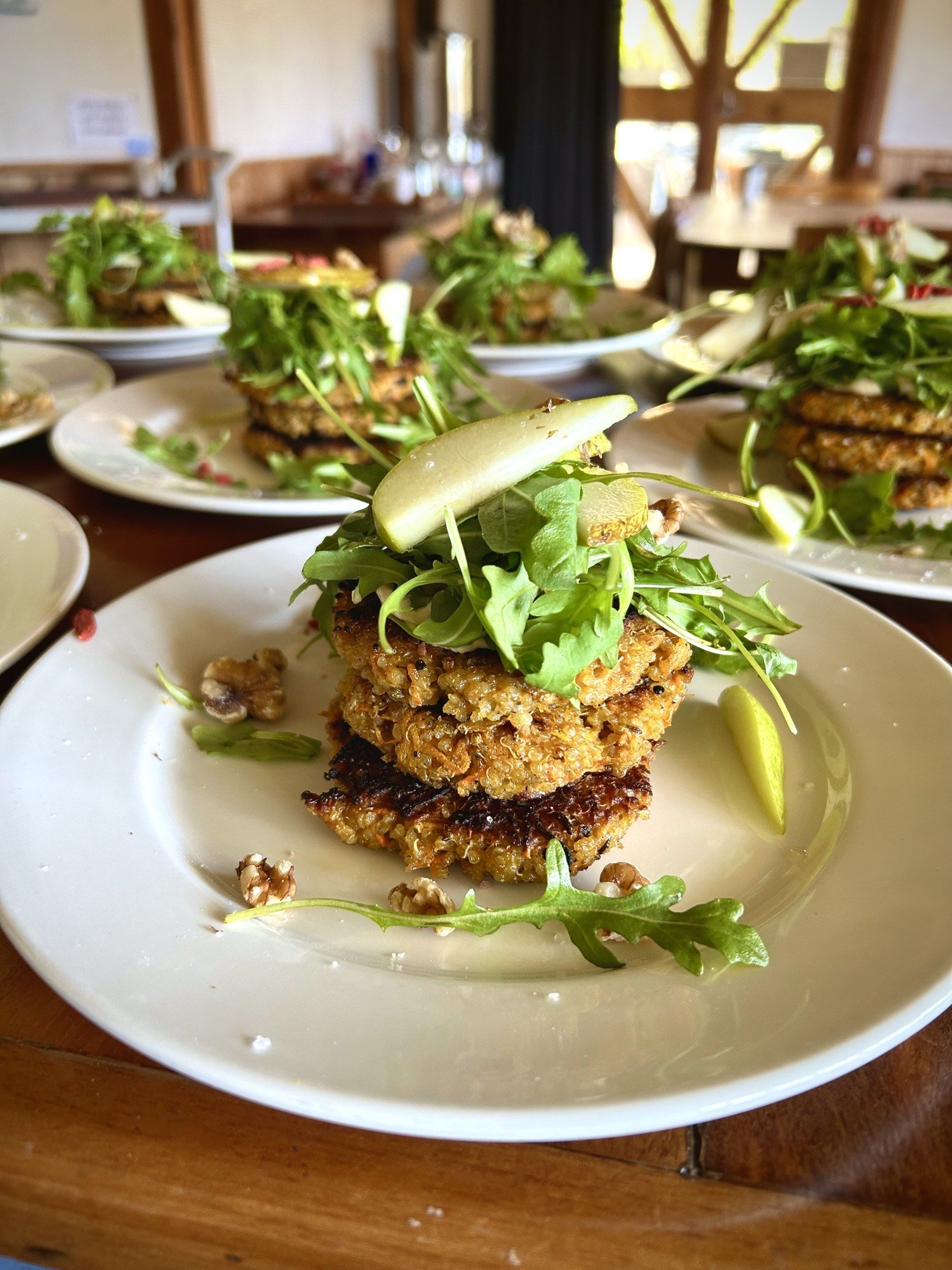 Retreat lunch meal of a plate of stacked vegan food topped with arugula, pickles, and microgreens, garnished with lime wedges, on a white plate with scattered walnut pieces, with more similar plates in the background inside a wooden dining area.