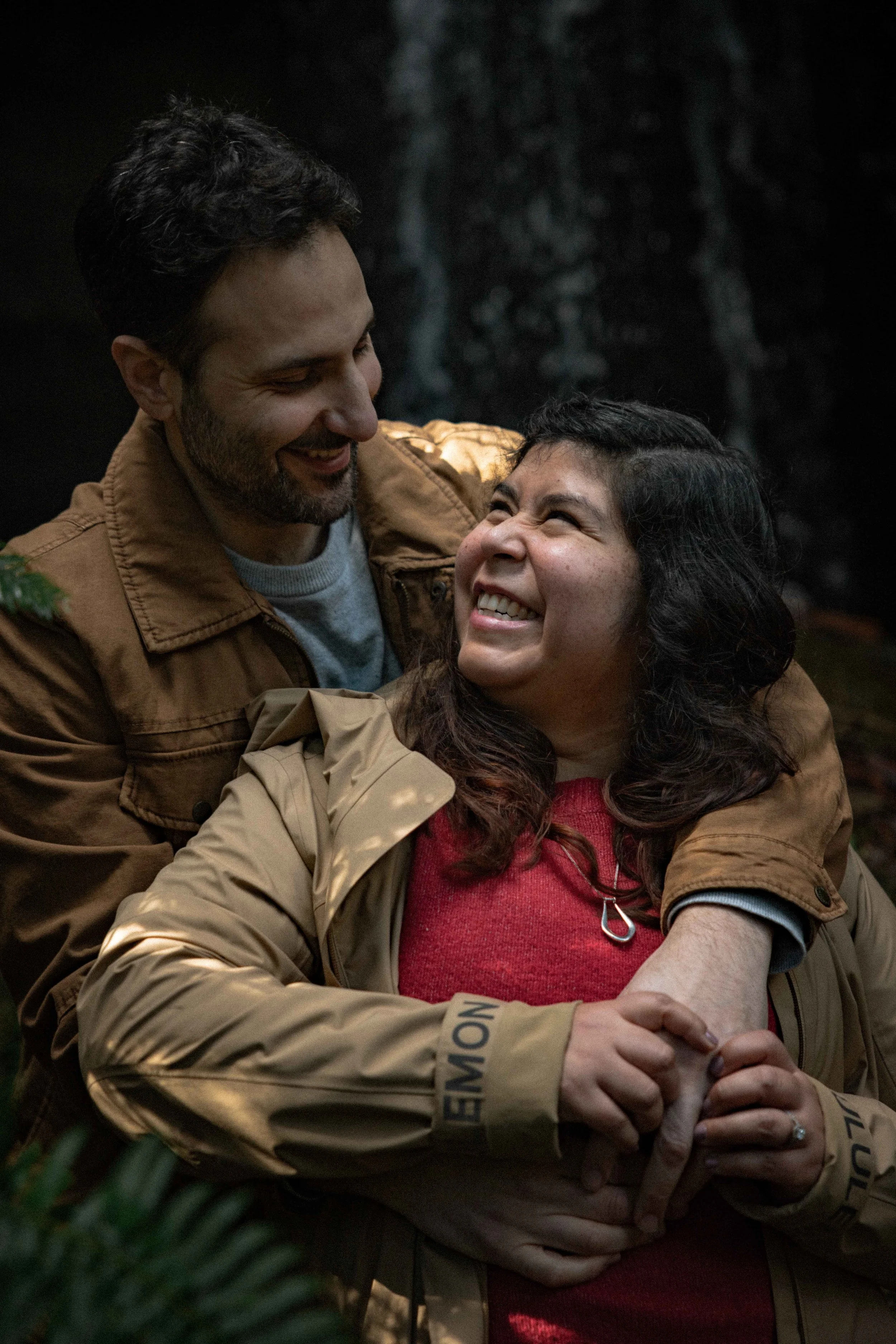 newly engaged couple look at each other  and smile while they hold each other in front of a waterfall in the forest