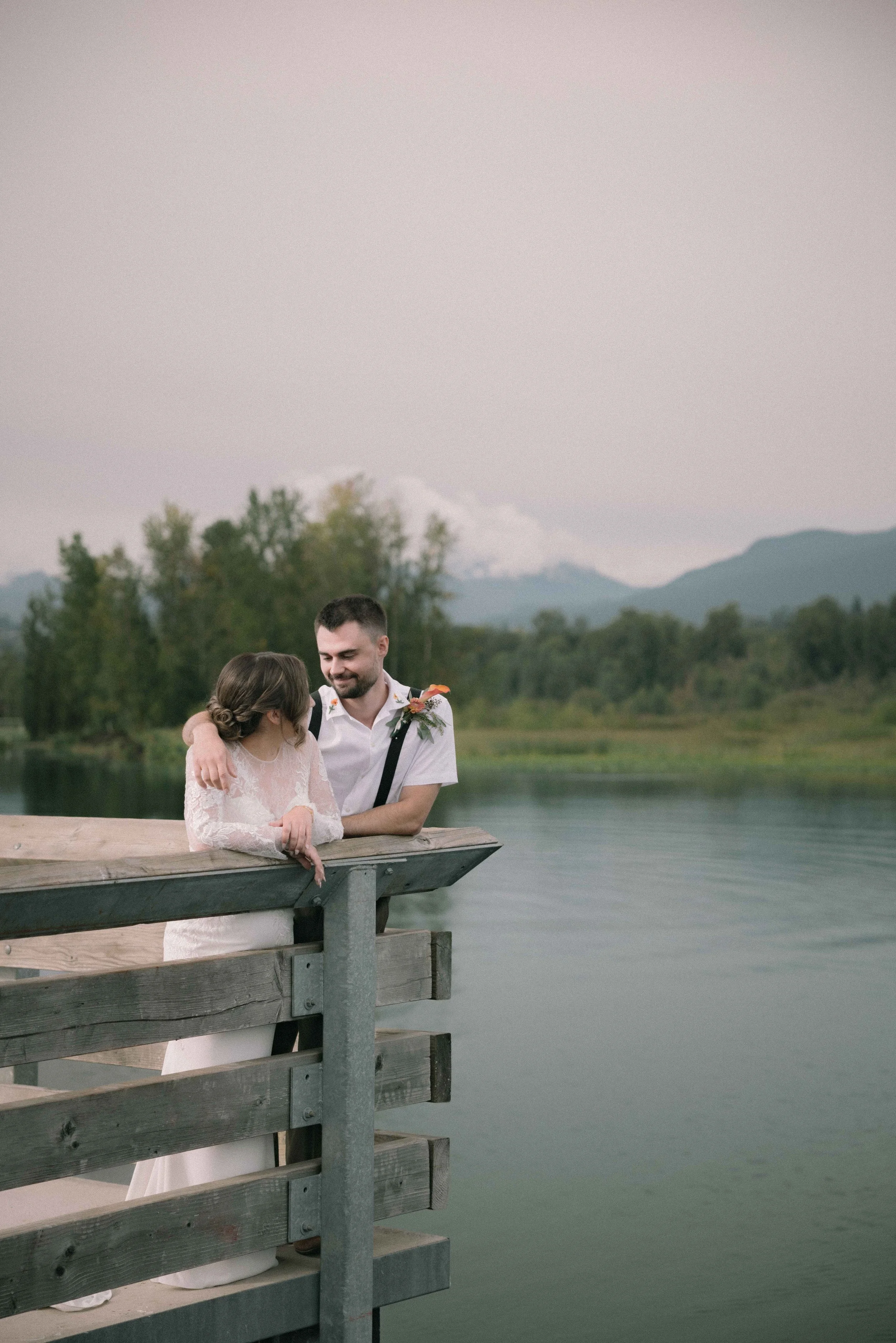 A bride and groom stand on the pier at the Burnaby Lake Pavilion with mountains in the background.