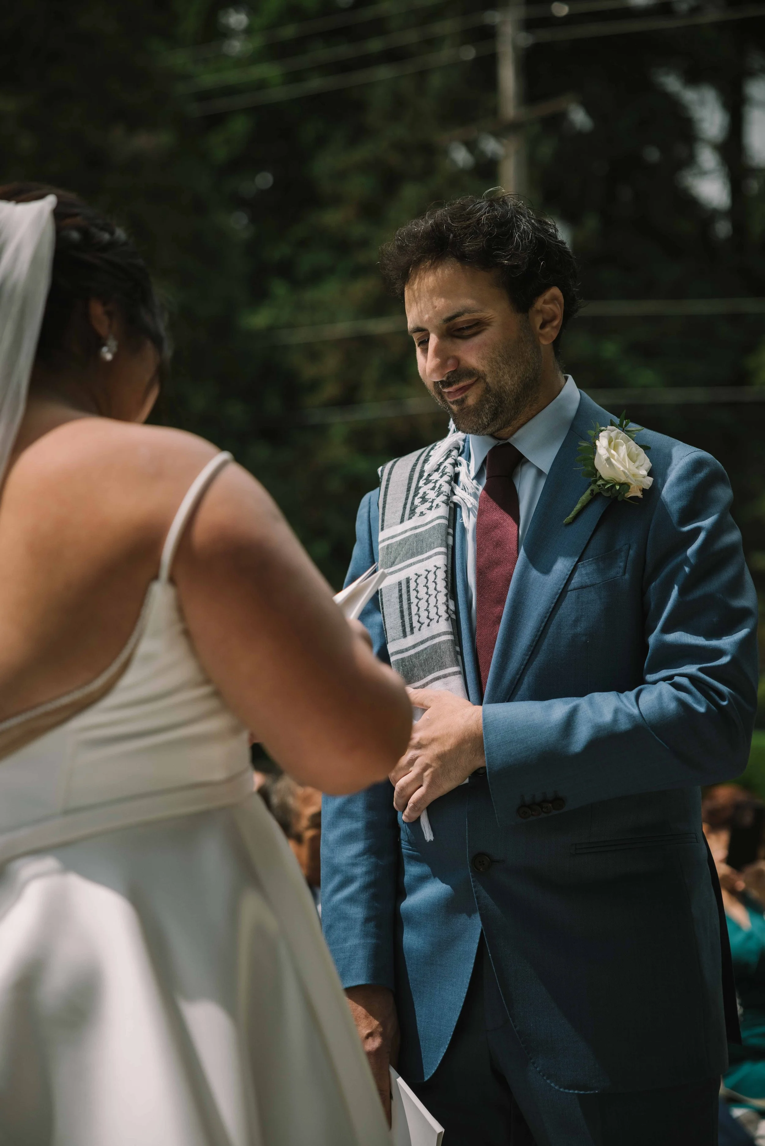 A groom wearing a blue suit with a traditional Lebanese scarf draped over one shoulder looks at his bride on their wedding day.