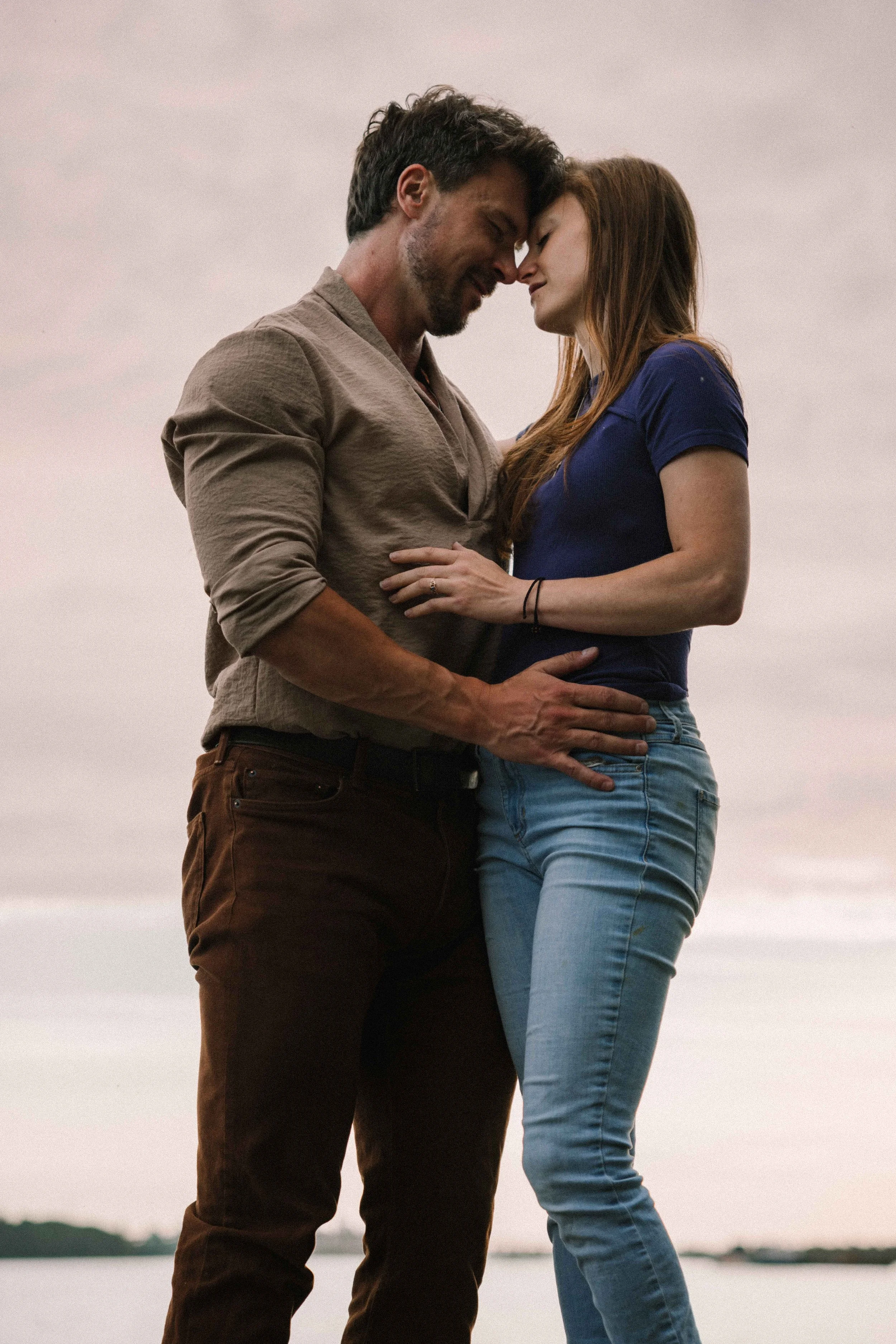 A man in brown pants and a linen shirt holds his fiancé waring blue jeans and a navy t shirt, with a pink and gold cloudy sky in the background on the beach. 