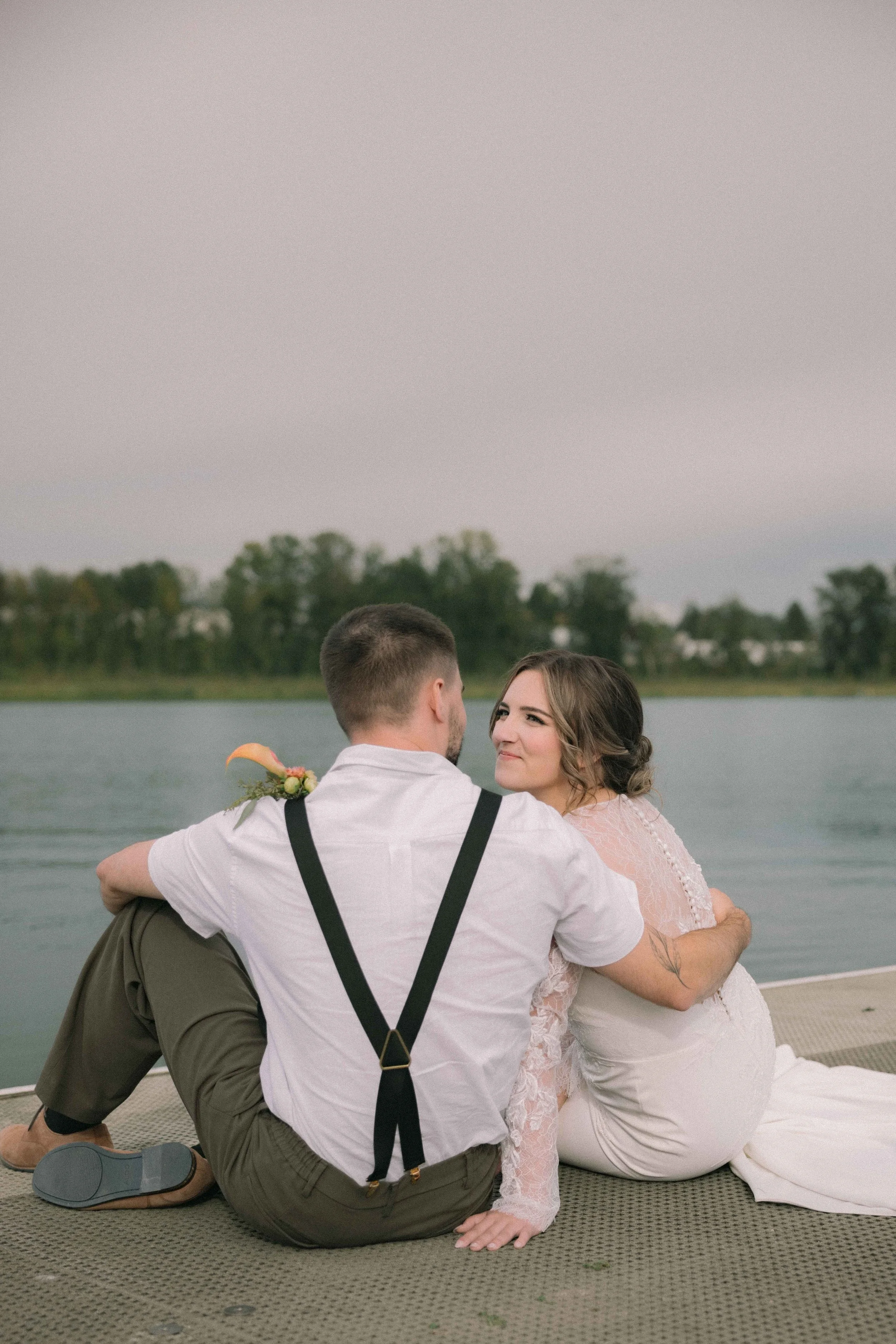 A bride sits on the edge of a dock at Burnaby Lake Pavilion looking at her groom on the waters edge