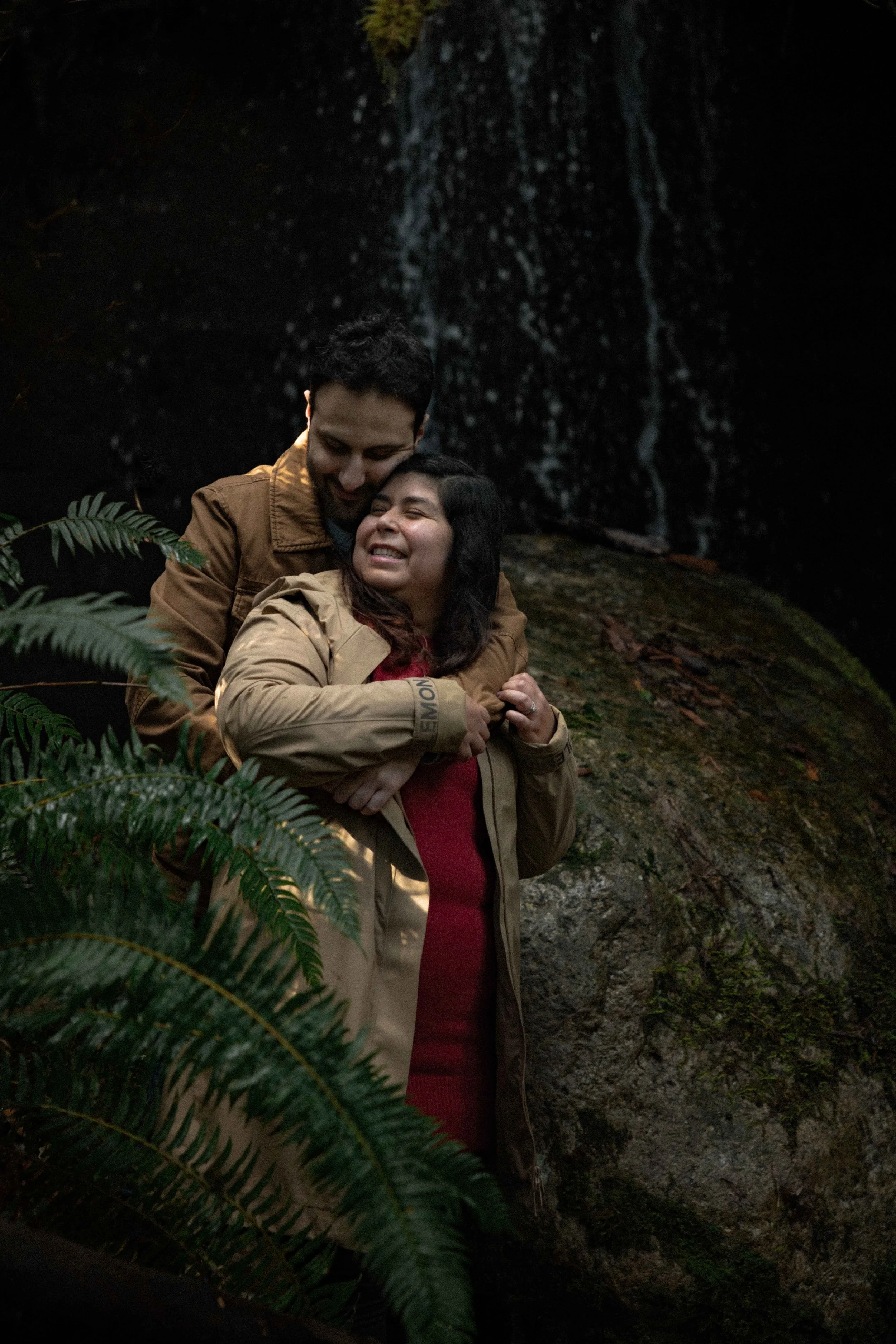 Man wearing tan coat hugs his fiancee from behind in front of a waterfall