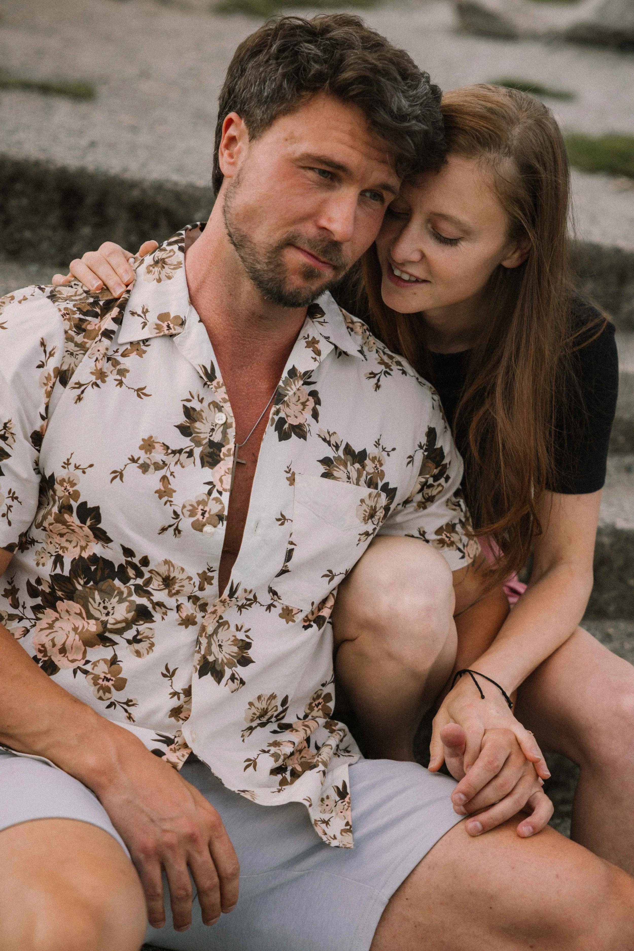 A newly engaged couple sit on stairs and look at the ocean as they lean into each other and cuddle.