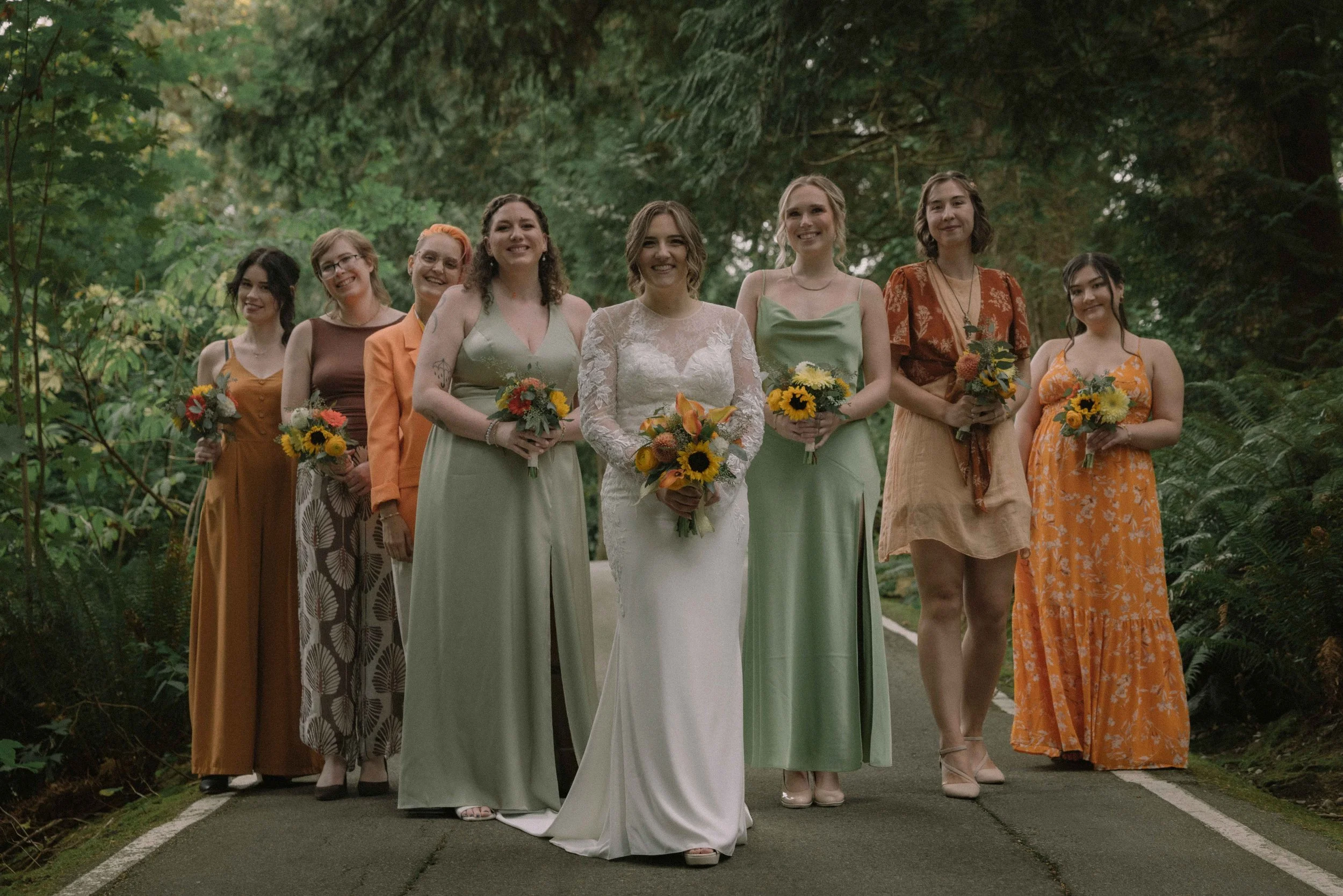 A bride with her bridal party wearing orange, soft ochre and sage green stands on the grounds of Minnekhada Lodge