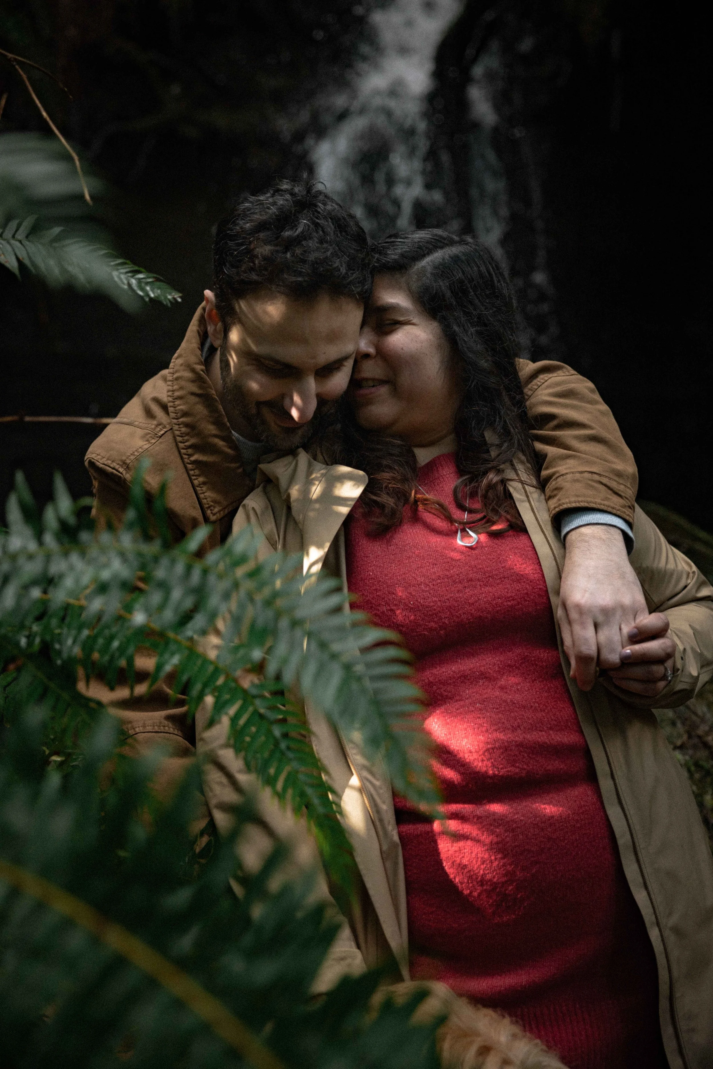 woman wearing a long trench coat nuzzles her fiancé in front of a waterfall in the forest