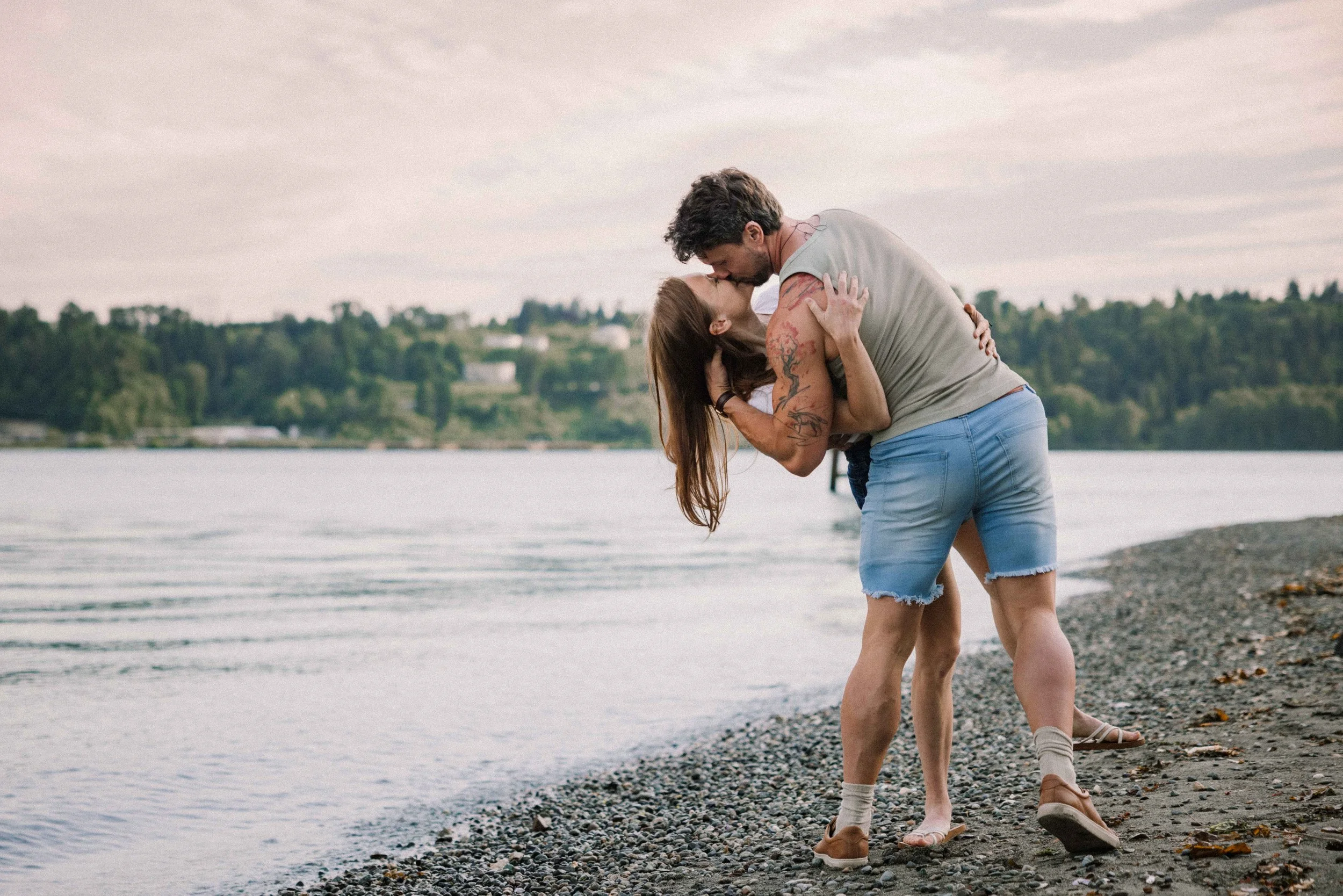 A man wearing blue jean shorts dips his fiance on the beach and kisses her.