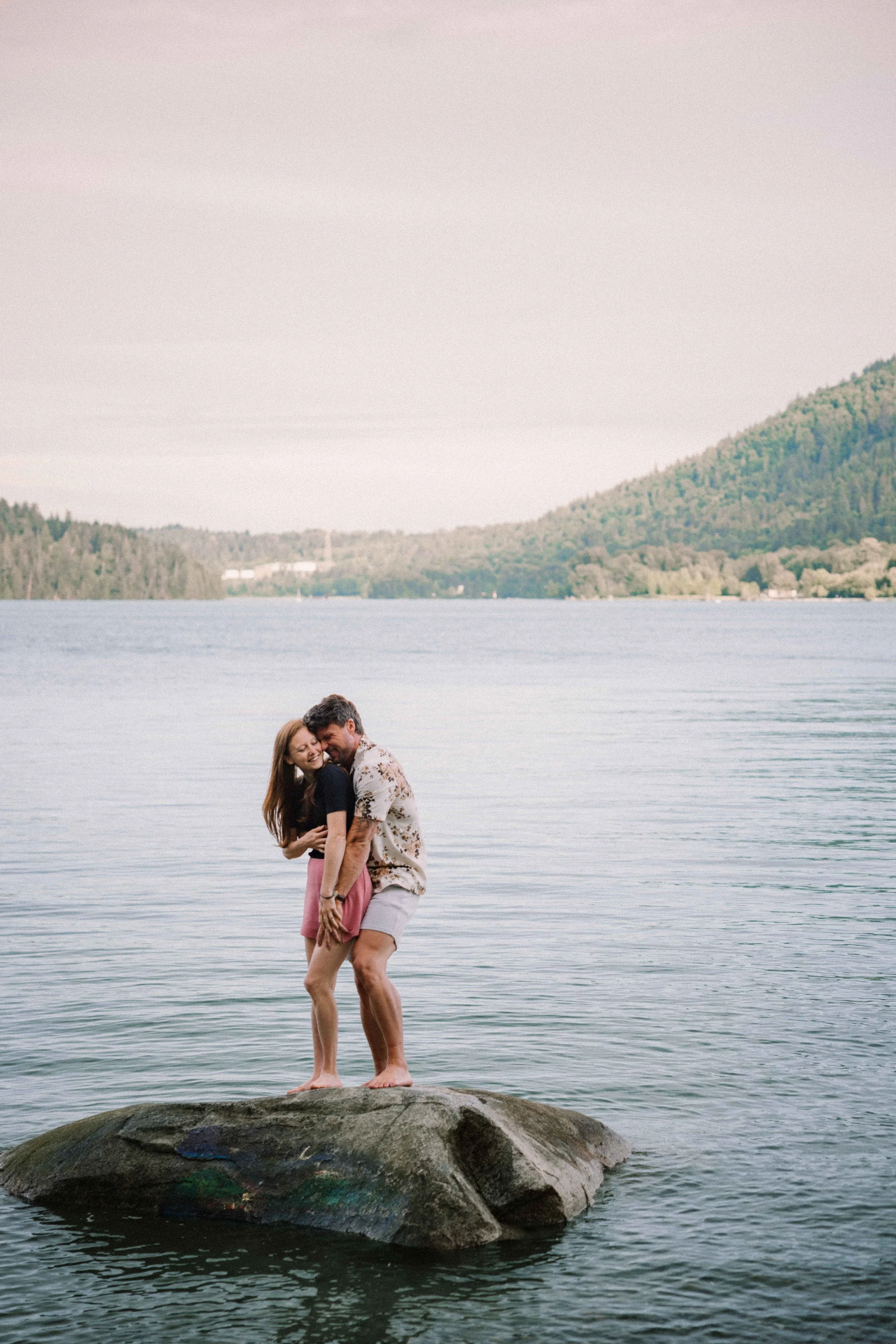 A red headed woman with pink shorts is hugged from behind by her future husband wearing a floral print shirt, as they stand on a rock in the middle of the ocean with mountains in the background.