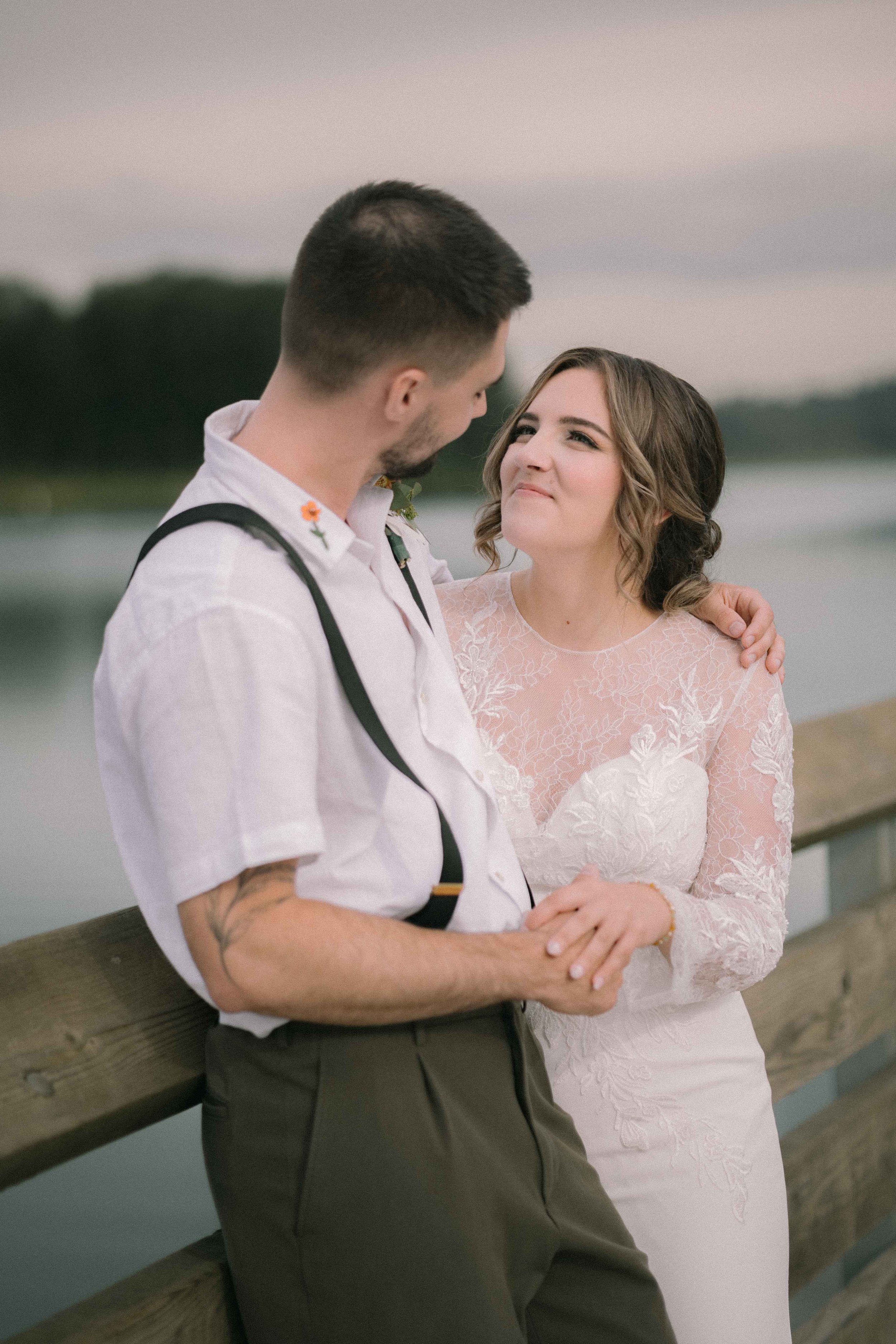 Newlyweds hold each other on the pier at Burnaby Lake Pavilion wearing an embroidered white linen shirt and a long sleeve lace dress. The bride has a textured low undo and natural makeup. 