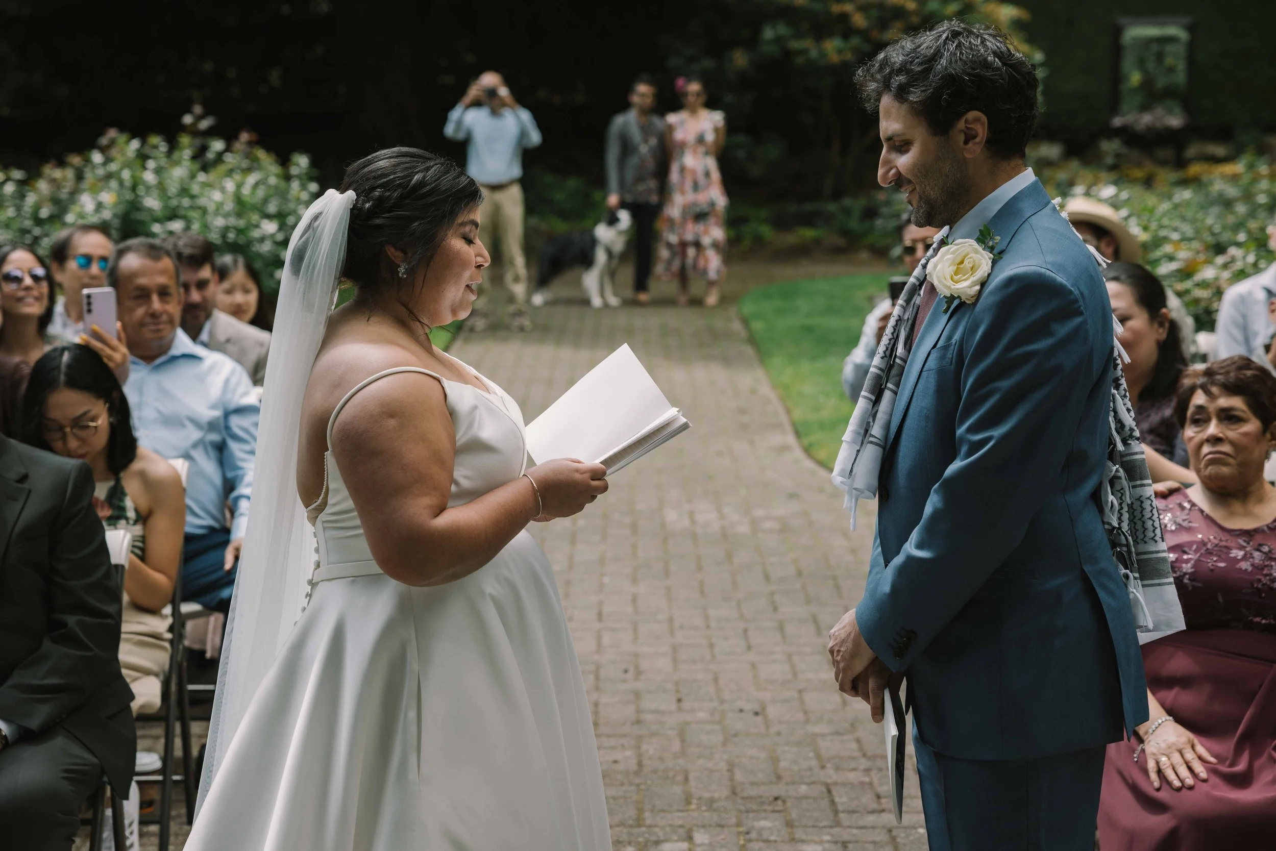 A bride with a veil in her low bun reads her vows to her groom at their wedding ceremony.