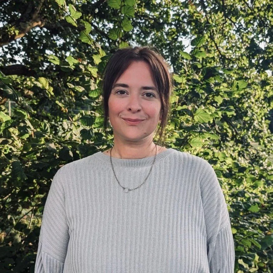 A woman with brown hair and fair skin standing outdoors in front of lush green foliage.