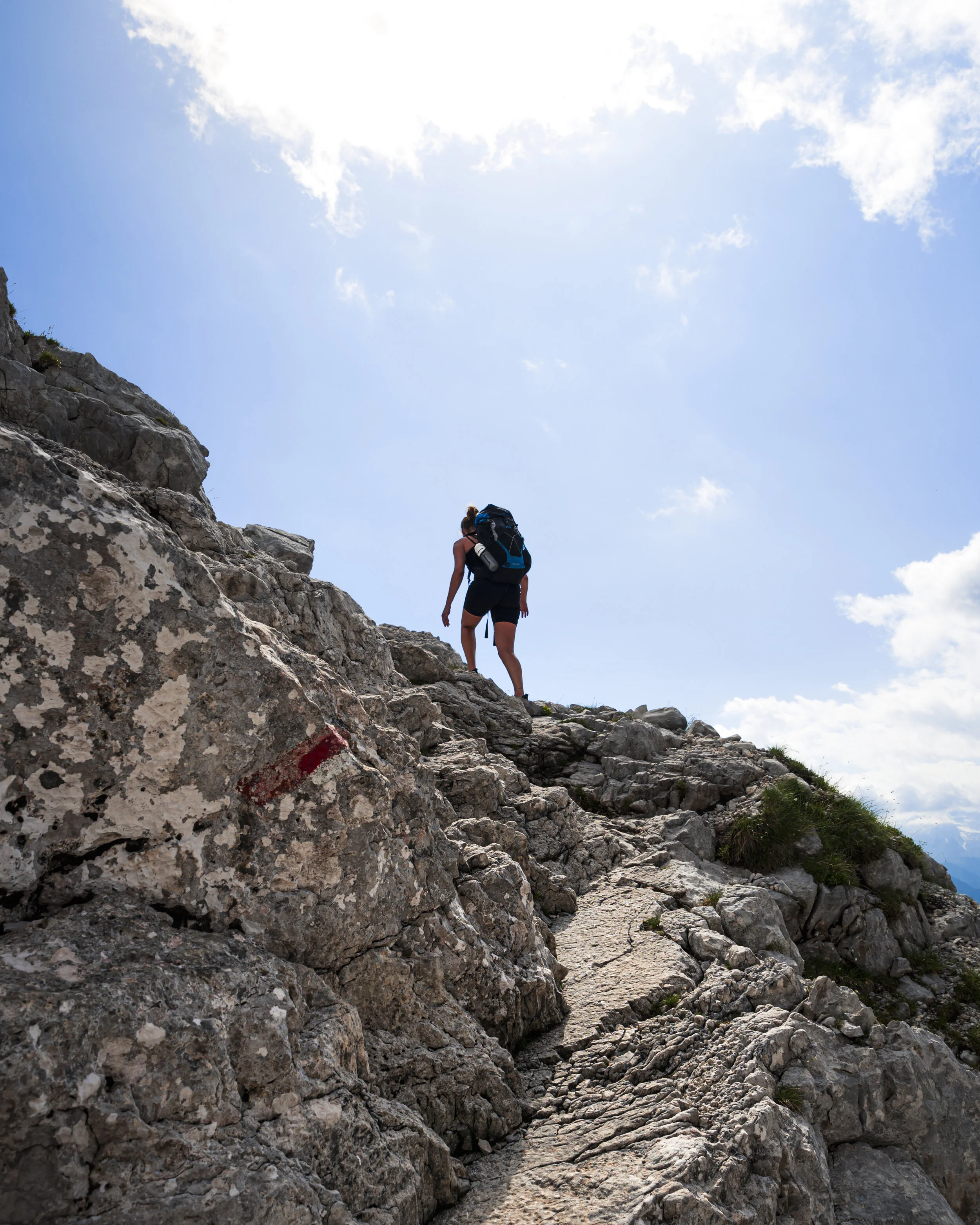 En person med rygsæk klatrer op ad en stejl, rocky bjergsti mod en blå himmel med nogle spredte skyer.