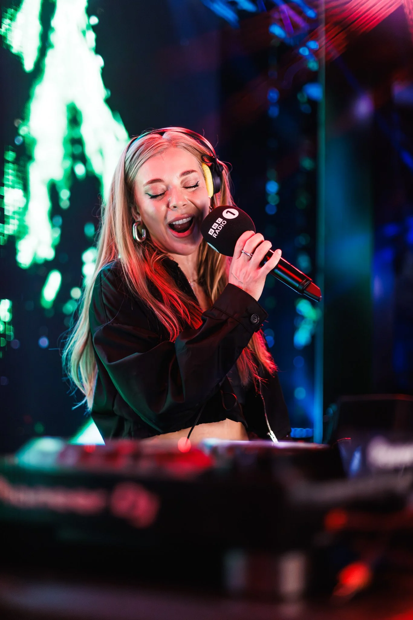 A female singer with blonde hair, wearing headphones and a black jacket, singing passionately into a microphone in a colorful, vibrant music studio or stage setting.