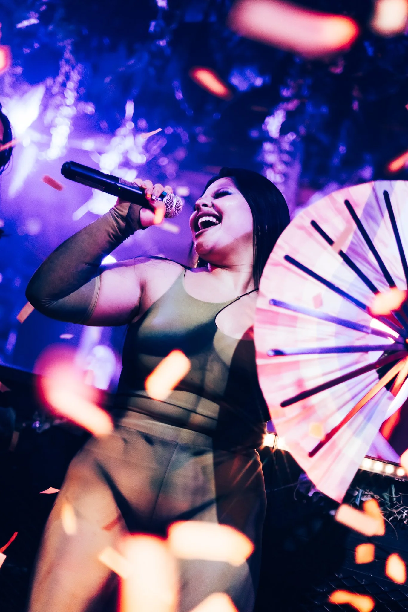 A woman singing with a microphone at a lively, colorful event surrounded by confetti and a decorative fan, under vibrant lighting.