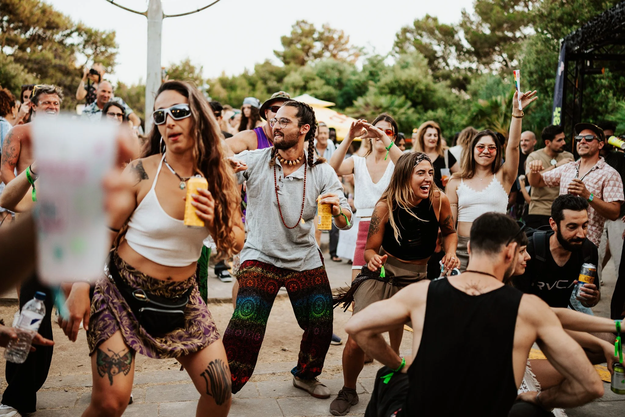 A lively crowd dancing and having fun outdoors at a festival or concert, with people of various ages and styles, surrounded by trees and tents.