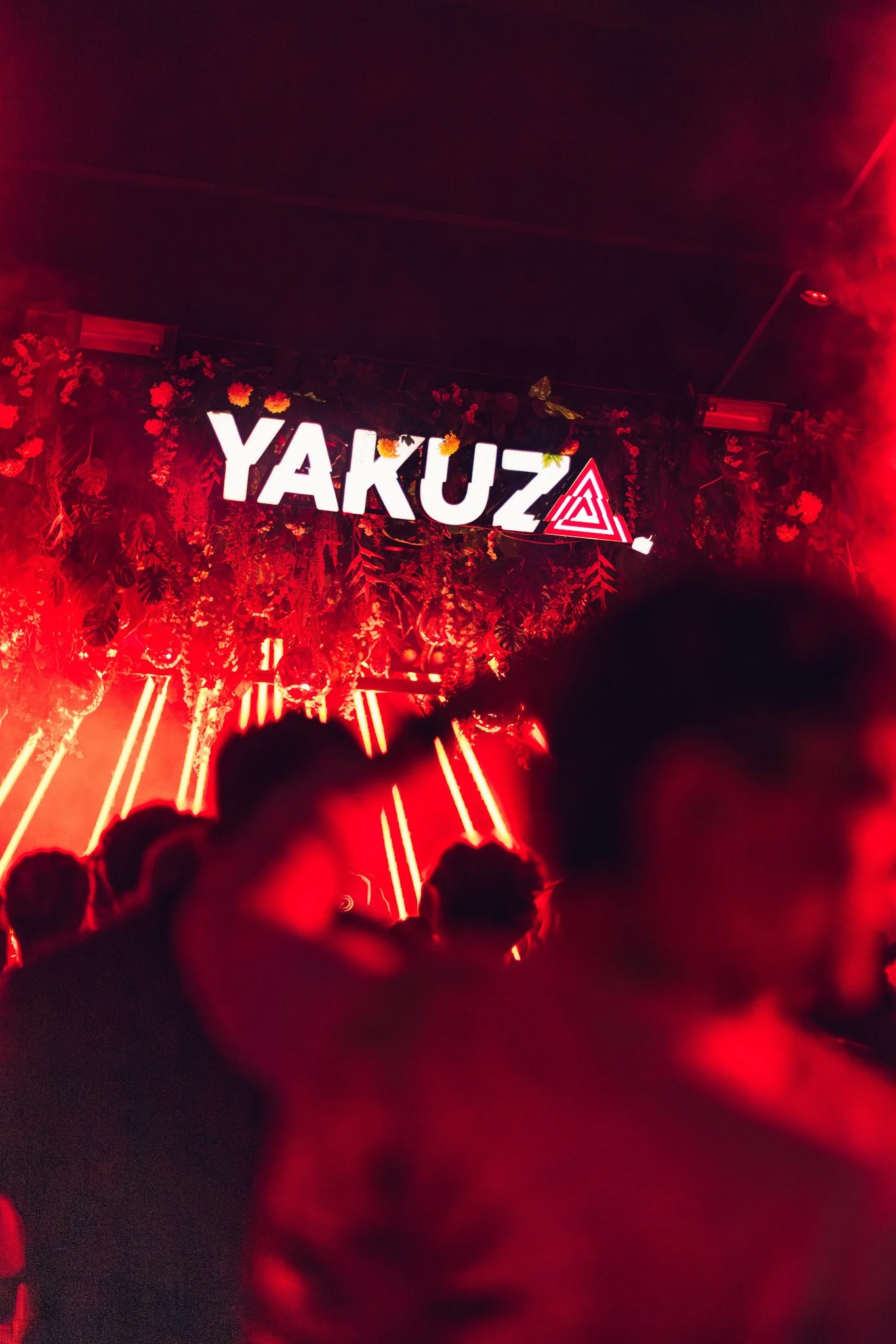 Red neon sign spelling 'YAKUZA' with floral decorations and red lighting, in a dark atmosphere, possibly a nightclub or bar interior.