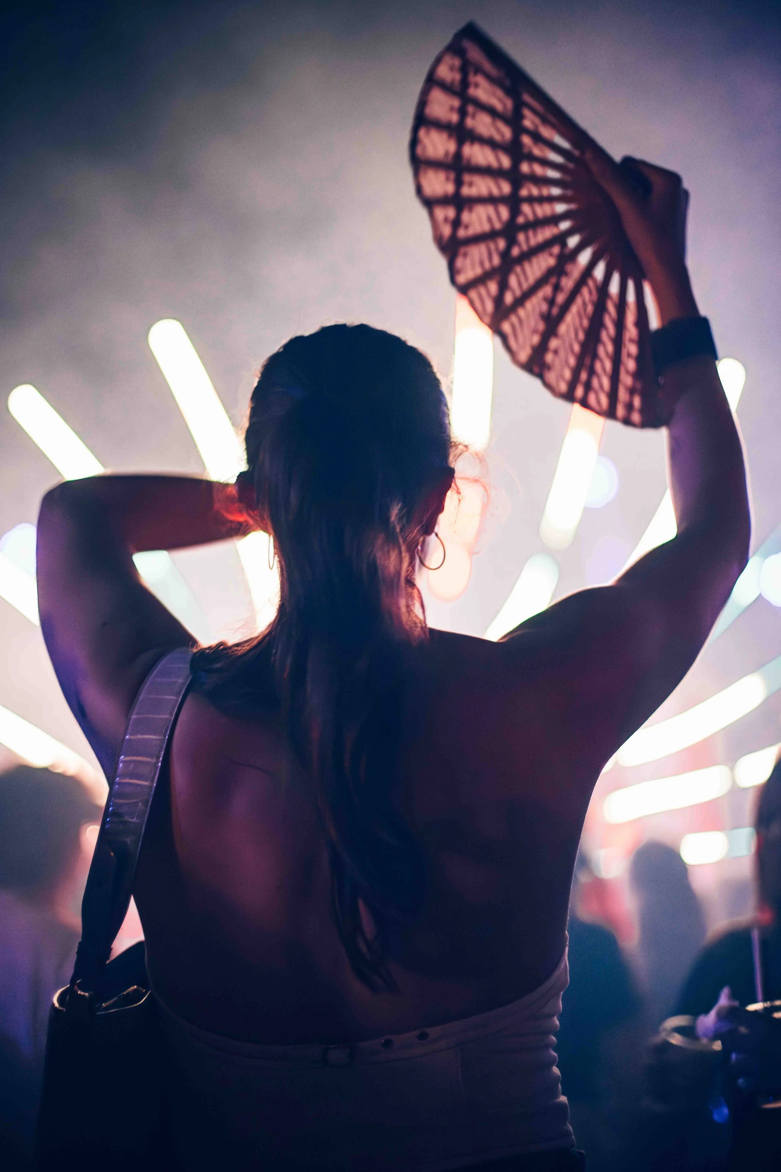 A silhouette of a woman holding a lace patterned fan above her head with her arms bent at the elbows in a nightclub or concert setting. There are colorful vertical lights in the background, creating a vibrant atmosphere.