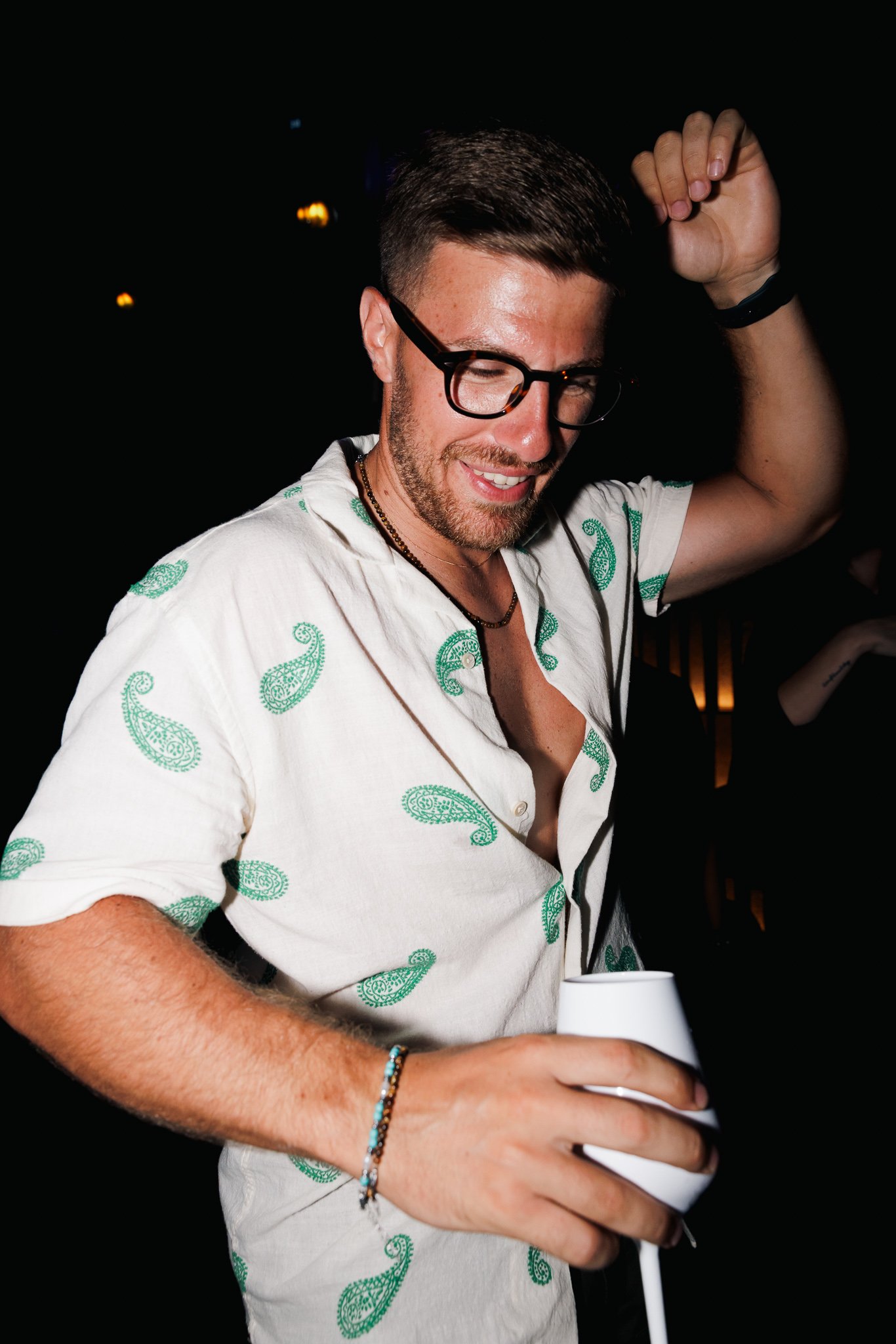 A young man with short dark hair, glasses, and a beard, smiling with eyes closed, raising his right arm, and holding a white cup in his left hand, wearing a white shirt with green paisley patterns, in a dark setting with some blurred lights in the ba