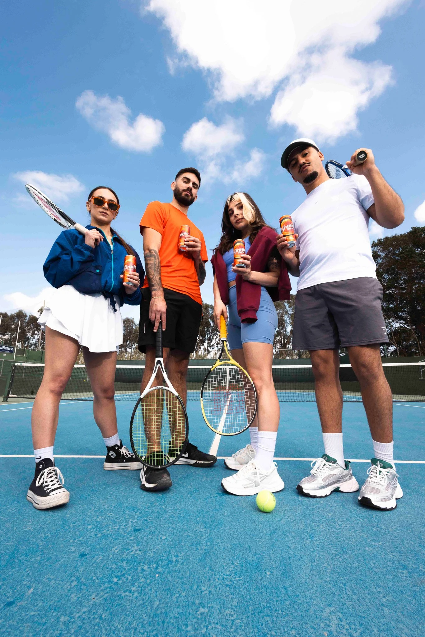 friends on a tennis court posing for the camera, holding rackets and kinnie cans in hand