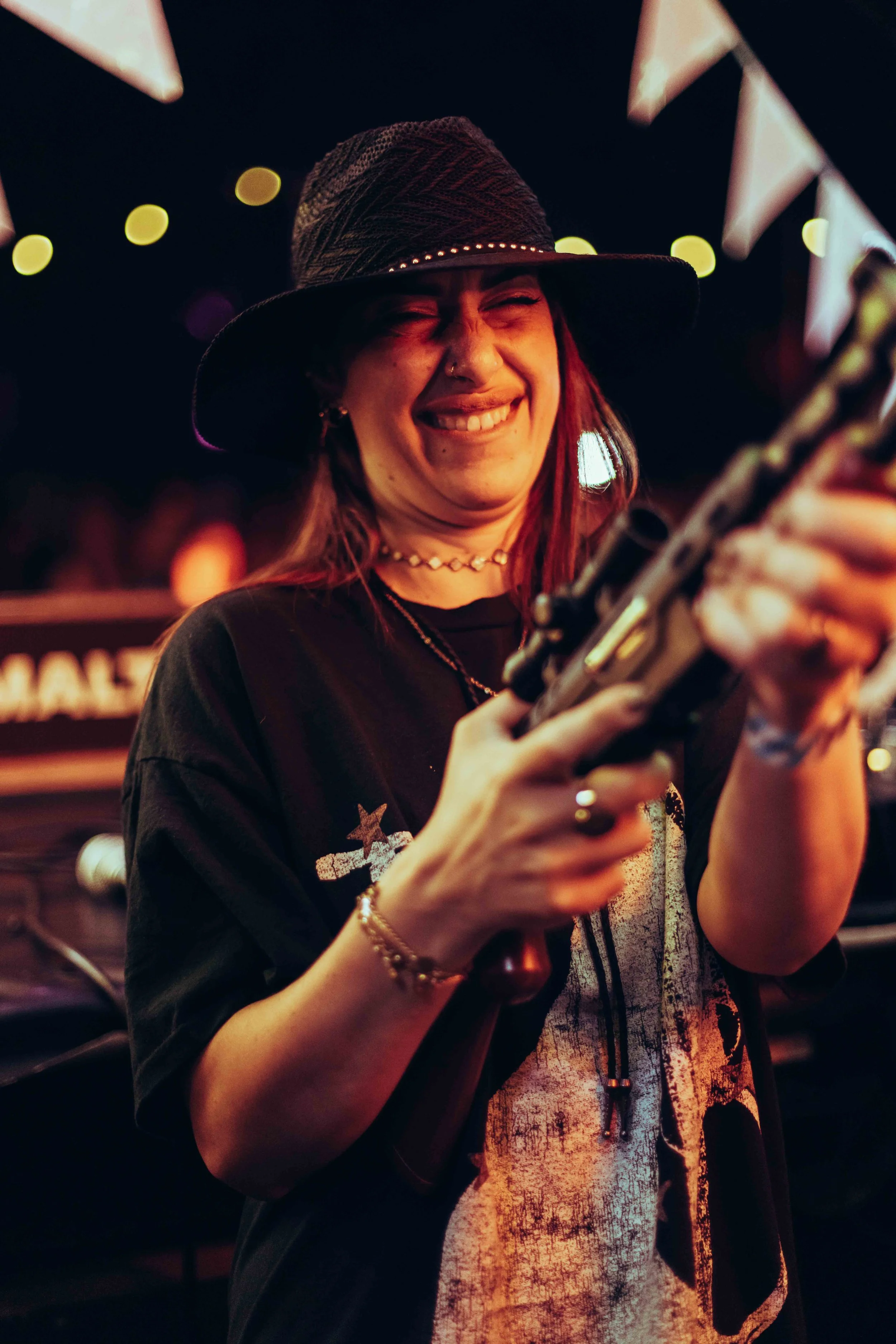 A young woman with red hair wearing a black hat with studs and a black t-shirt with stars, smiling while holding a microphone at night with colorful lights in the background.