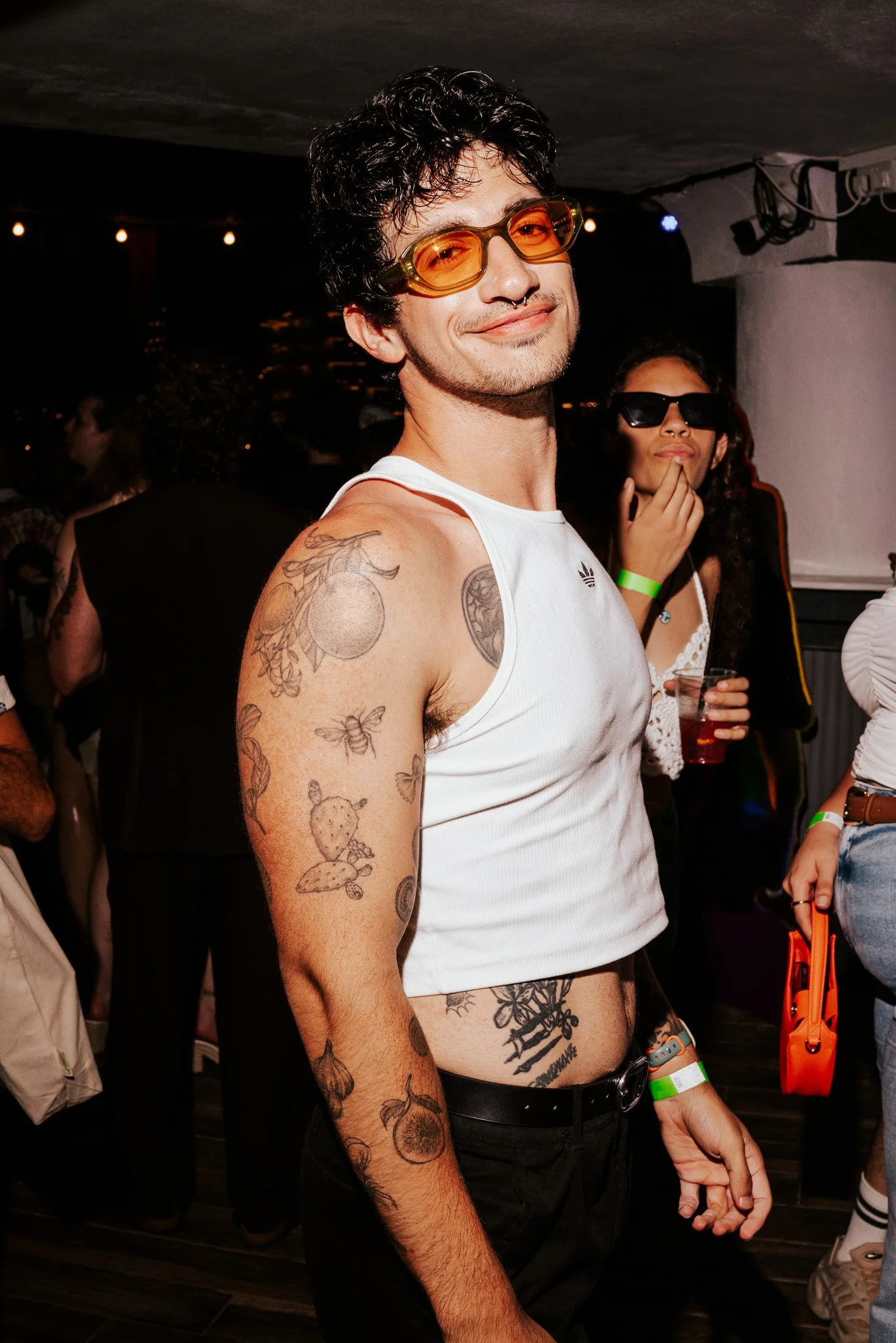 A young man with dark curly hair and tattoos on his arms and abdomen, wearing orange-tinted sunglasses, a white tank top, and a green wristband at a lively indoor event. He is smiling and looking at the camera, with a woman wearing dark sunglasses an