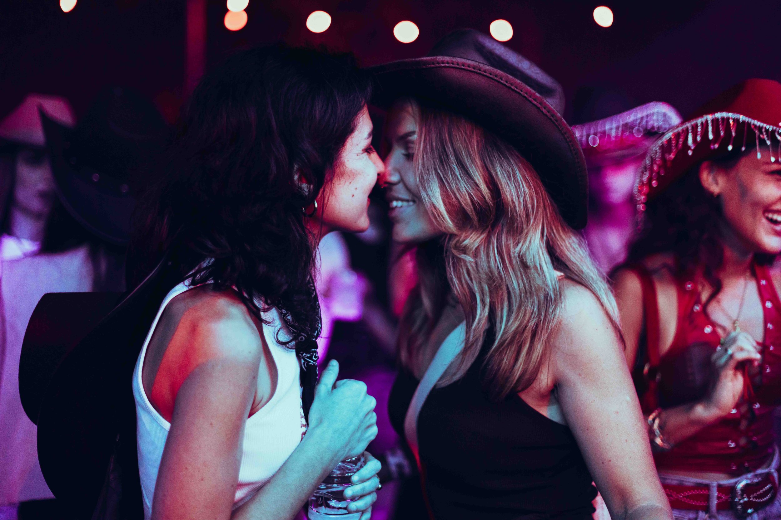 Two women dressed as cowgirls about to kiss at a costume party or themed event, with a third woman in a similar costume in the background.
