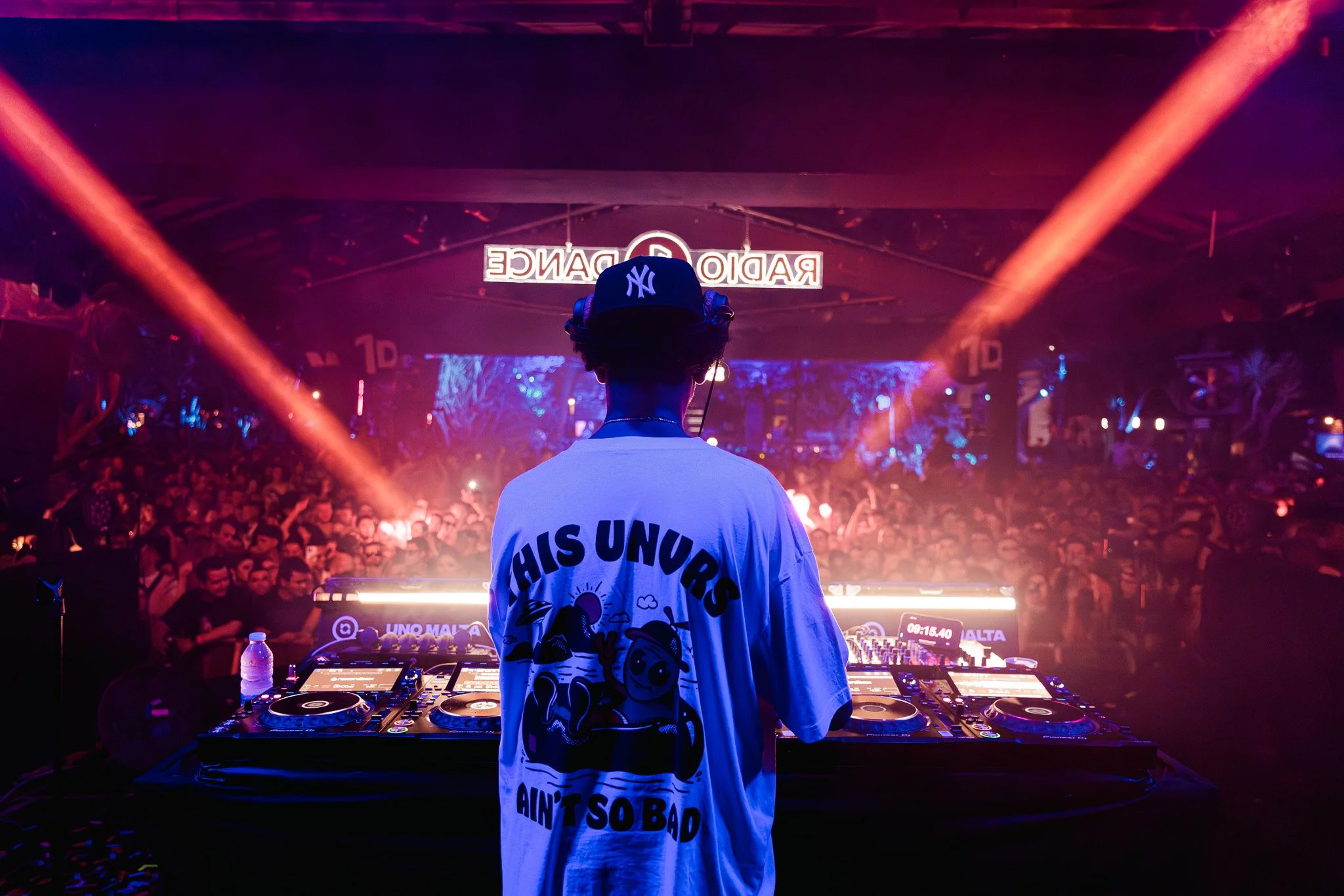 DJ wearing a cap with New York Yankees logo and a shirt with the text 'HIS UNORS AIN'T SO BAD' standing behind a DJ booth at a crowded nightclub with colorful stage lights and a large crowd in the background.