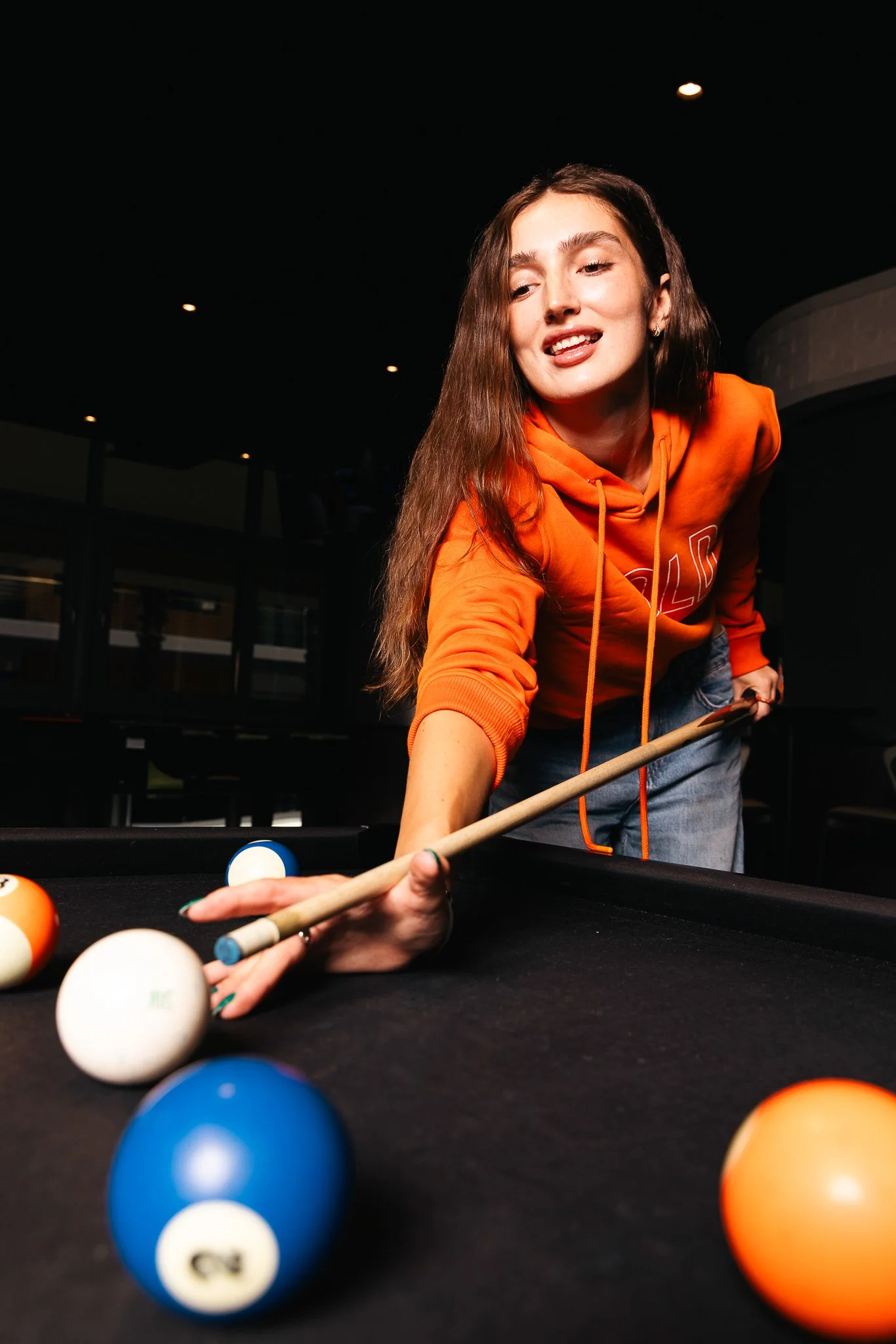 woman about to strike her shot on a billiard table, wearing an orange hoodie, kinnie winter shoot