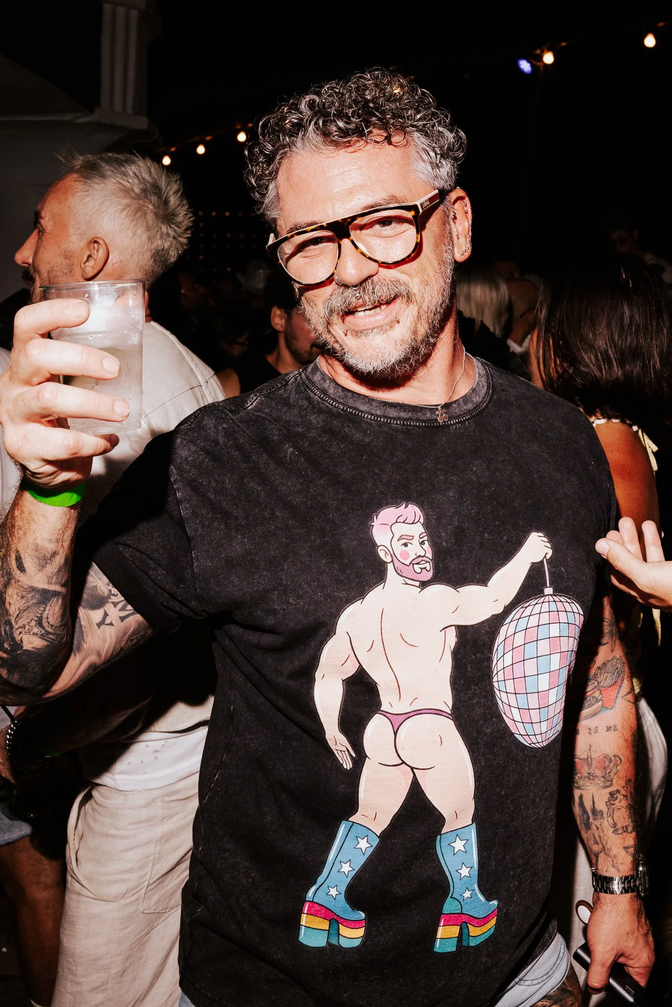 A man with curly gray hair, glasses, and a beard, smiling at a party. He's wearing a black T-shirt with a cartoon graphic of a muscular person in roller skates, holding a disco ball. Other people are visible in the background, some holding drinks.