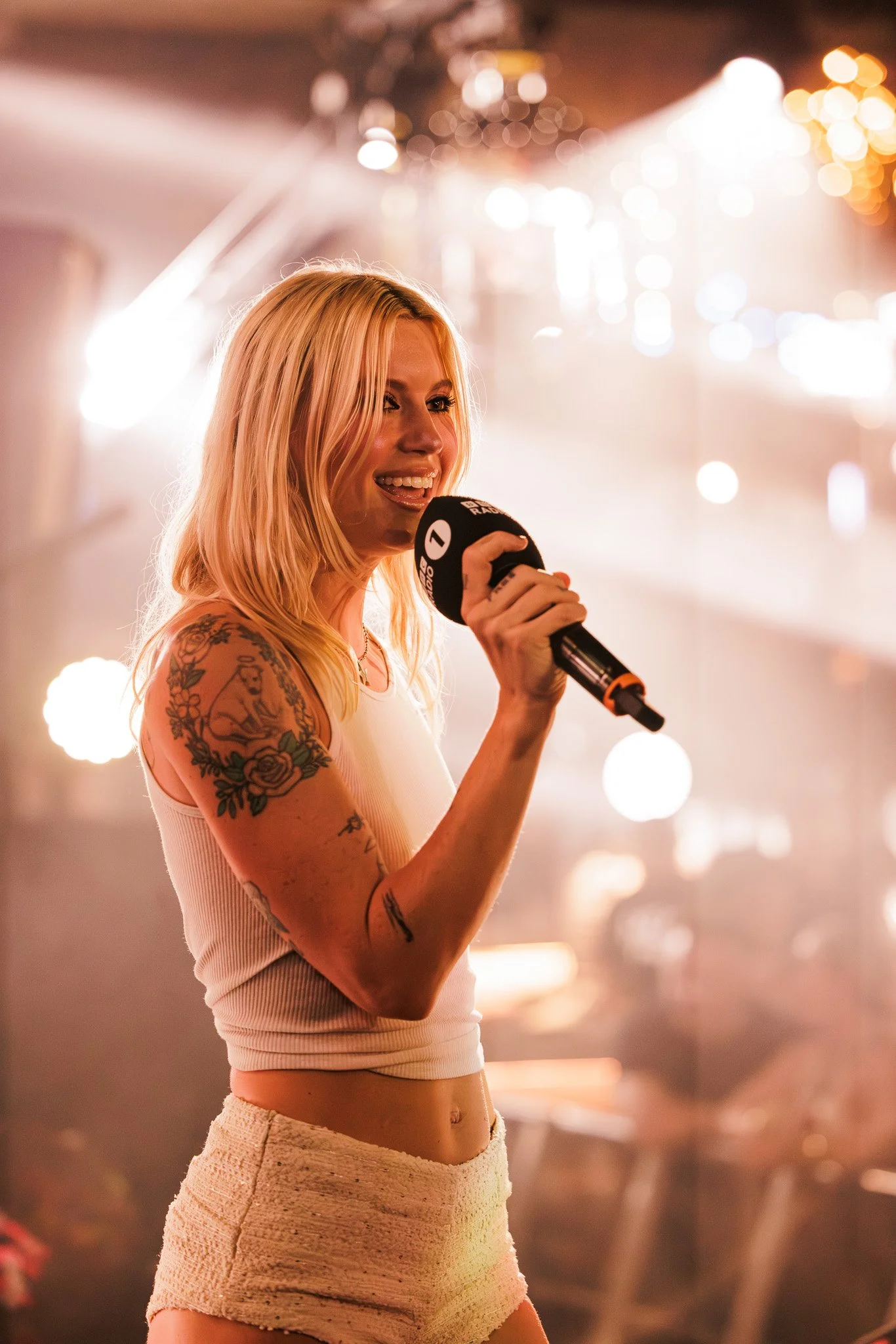 A young woman with blonde hair and tattoos on her arms, smiling and speaking into a microphone at an indoor event with bokeh lights in the background.