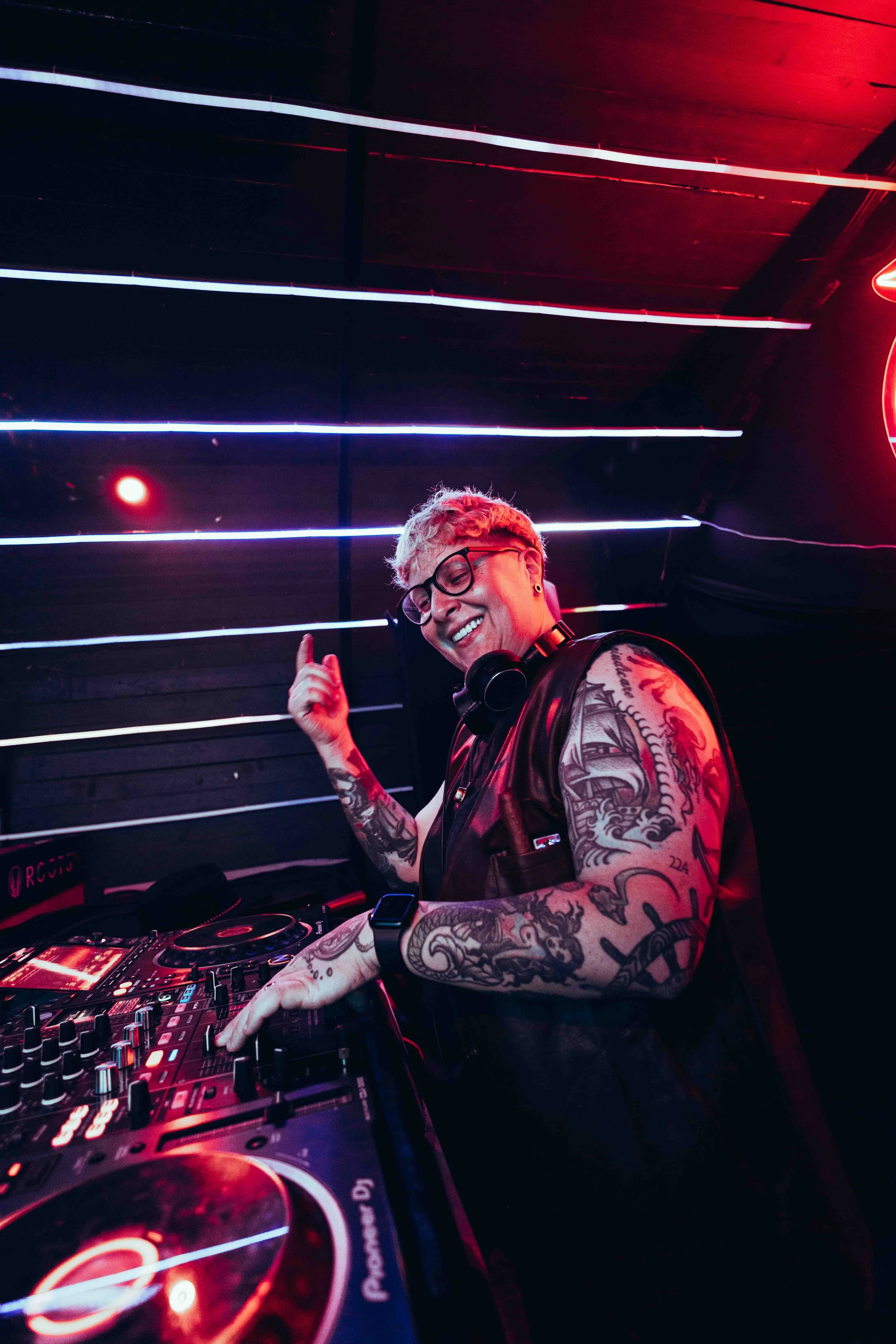 A smiling DJ with tattoos and glasses, wearing headphones, pointing upward while mixing tracks at a DJ booth in a club with colorful LED lighting.