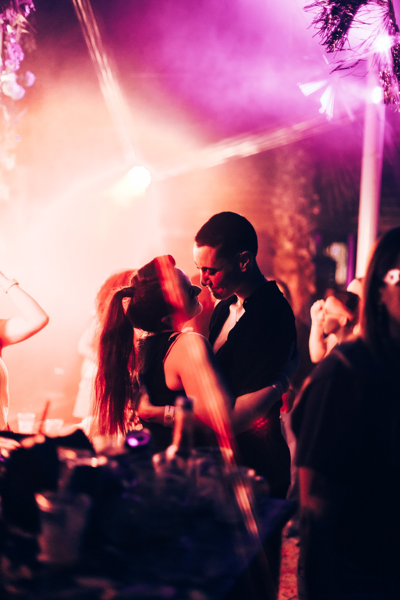 Two people are dancing closely together in a dimly lit, colorful nightclub with pink and purple lights and palm tree decorations in the background.