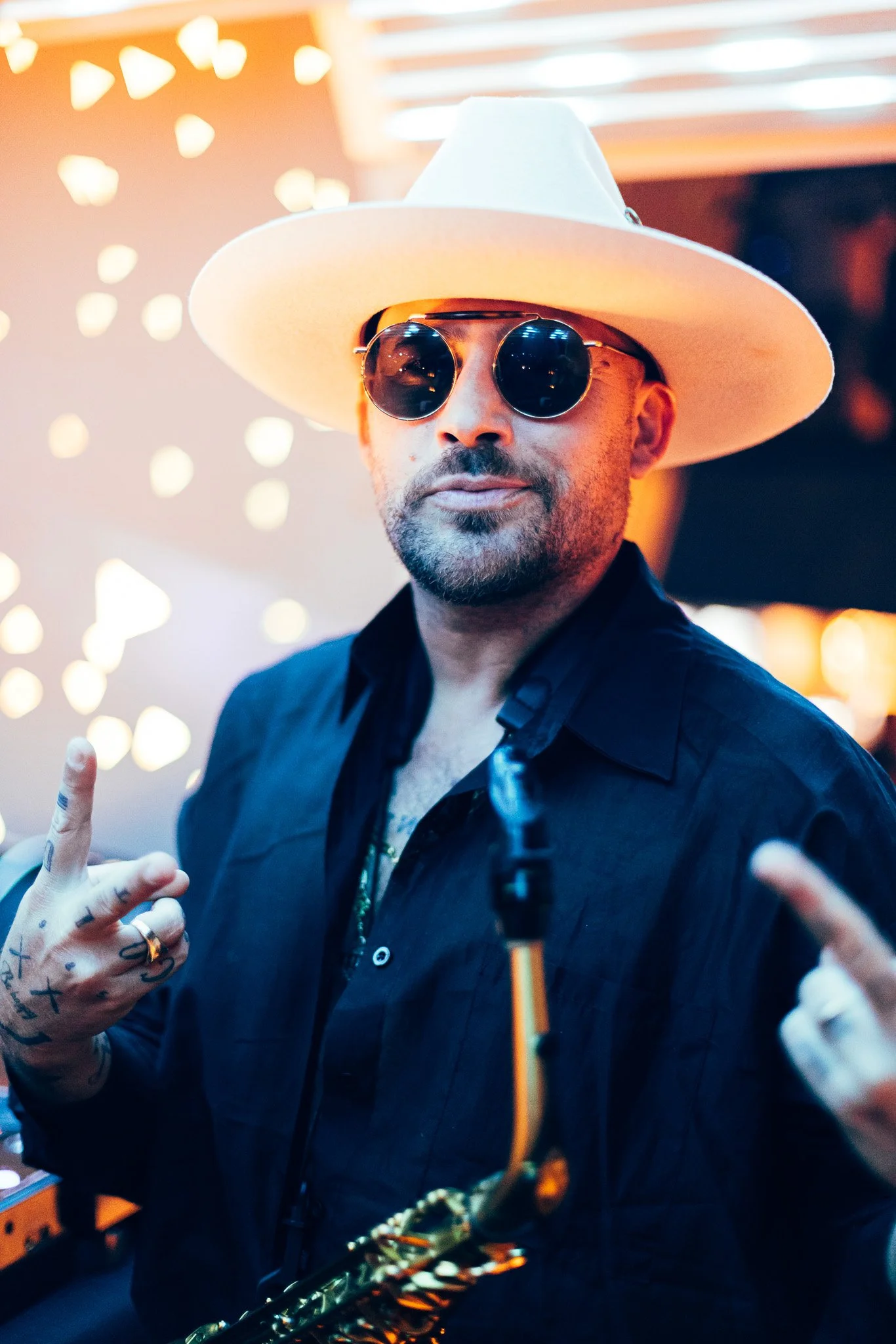 A man wearing a white wide-brimmed hat, round sunglasses, and a dark shirt, making a peace sign with his tattooed hand, standing in a dimly lit setting with warm, blurry lights in the background.